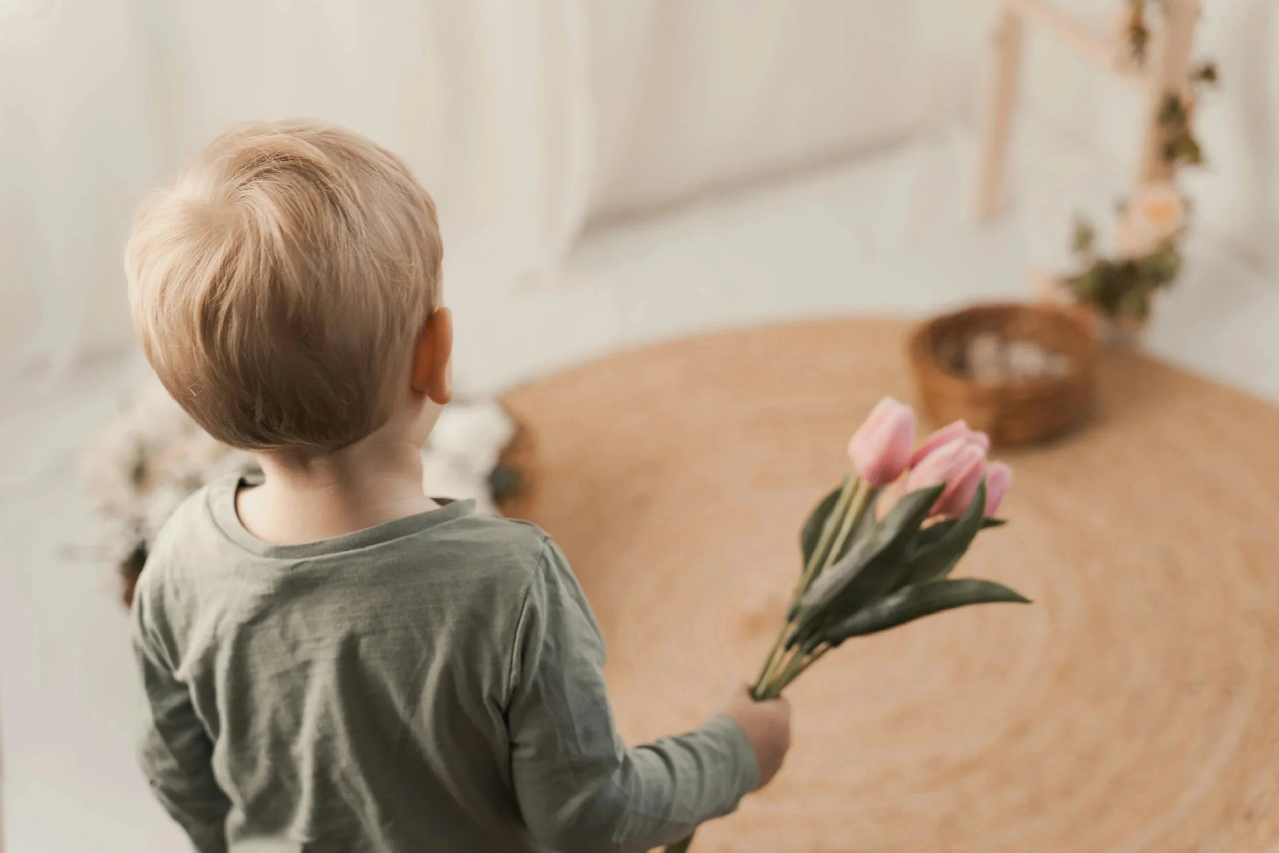 A young boy with blonde hair holding a small bouquet of pink tulips, standing near a wooden table with a basket and a floral arrangement in the background.