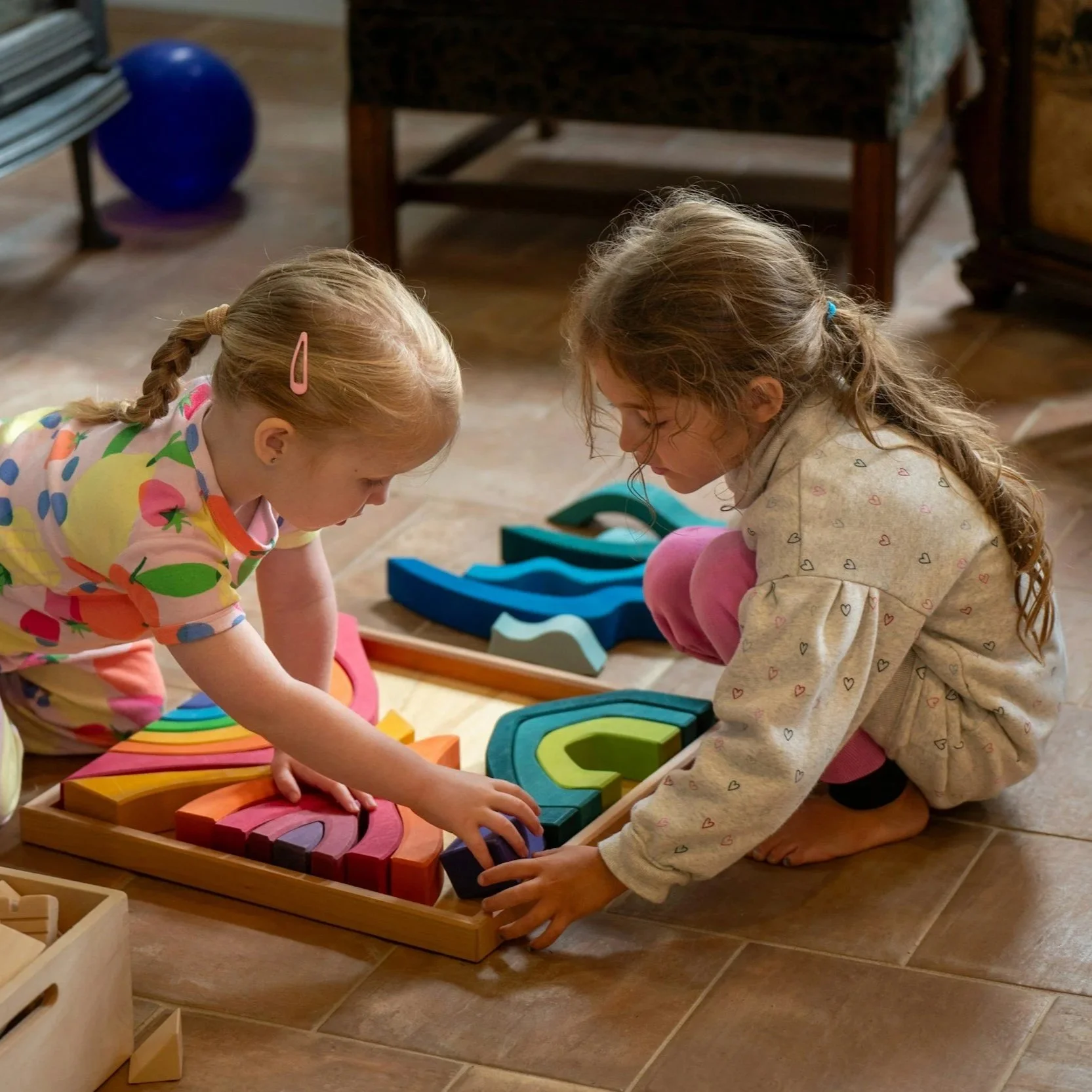 Two young girls playing with colorful wooden rainbow and block toys on the floor in a cozy room.