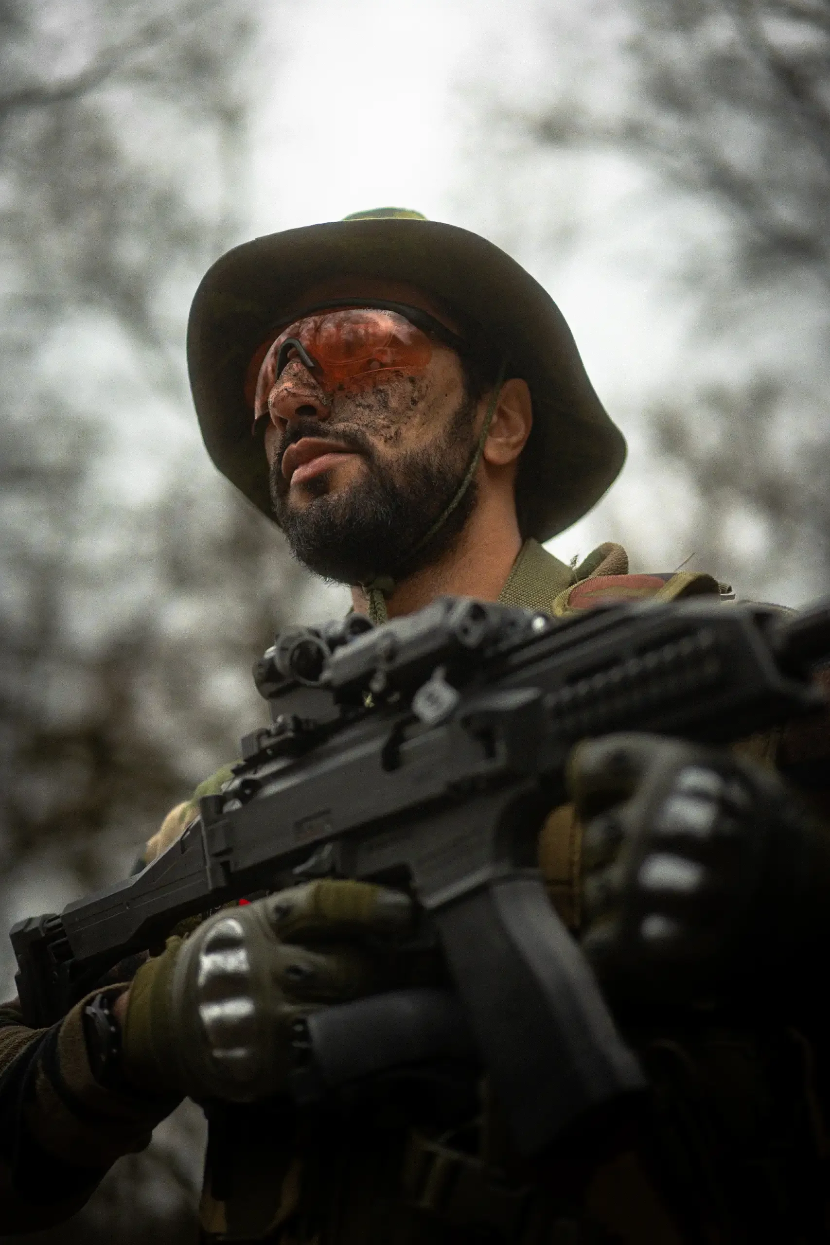 Un soldat avec un casque, lunettes de soleil, gilet tactique et arme à feu, dans un environnement extérieur. La lumière est diffuse et le fond est flou.