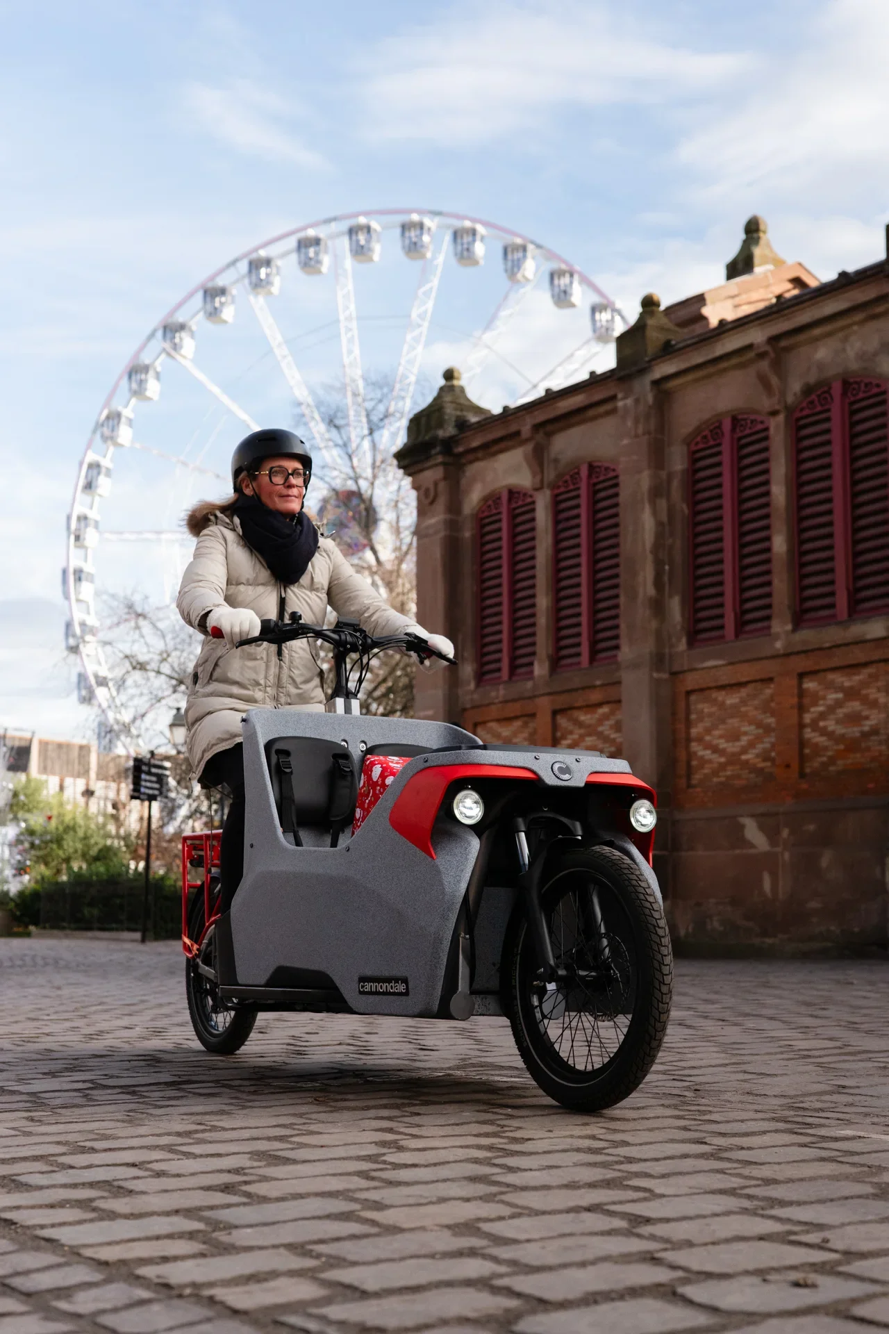 Une femme en armure de moto et casque, roulant sur un vélo électrique dans une rue pavée, avec une grande roue de fête foraine en arrière-plan.