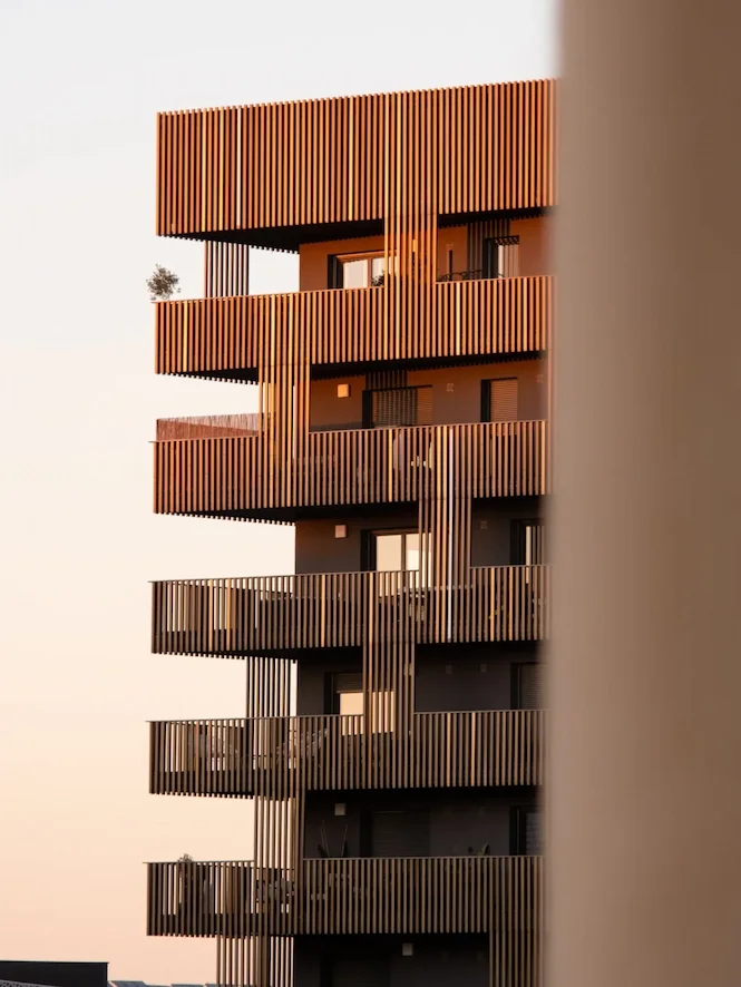 Un bâtiment résidentiel moderne avec plusieurs étages, chaque étage possède un balcon en bois aligné verticalement, éclairé par la lumière du coucher de soleil, avec un mur partiellement visible sur le côté droit de l'image.