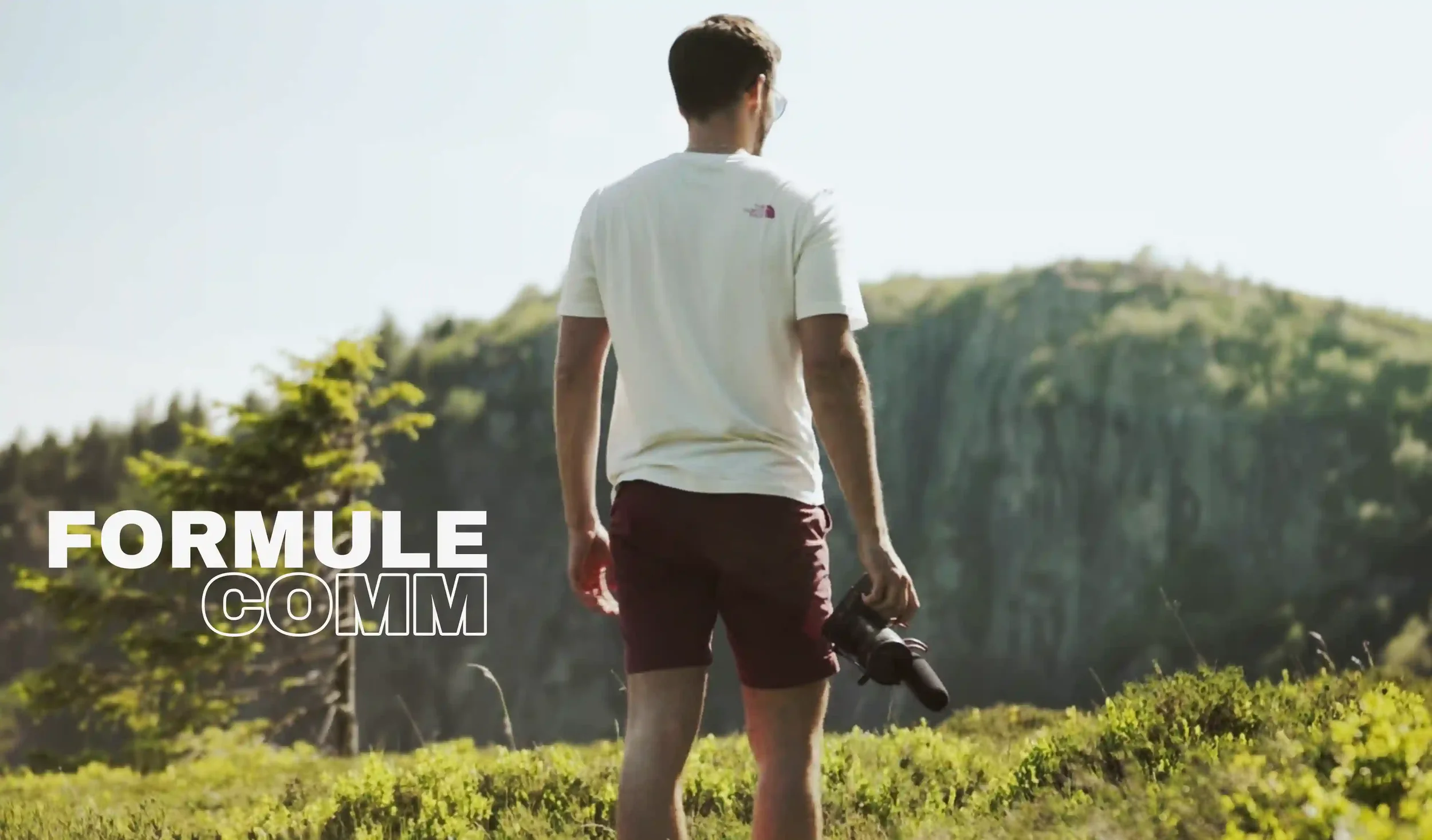 Un homme marche dans un paysage nature, tenant un trépied avec une montagne en arrière-plan, avec le texte "Formule COM".