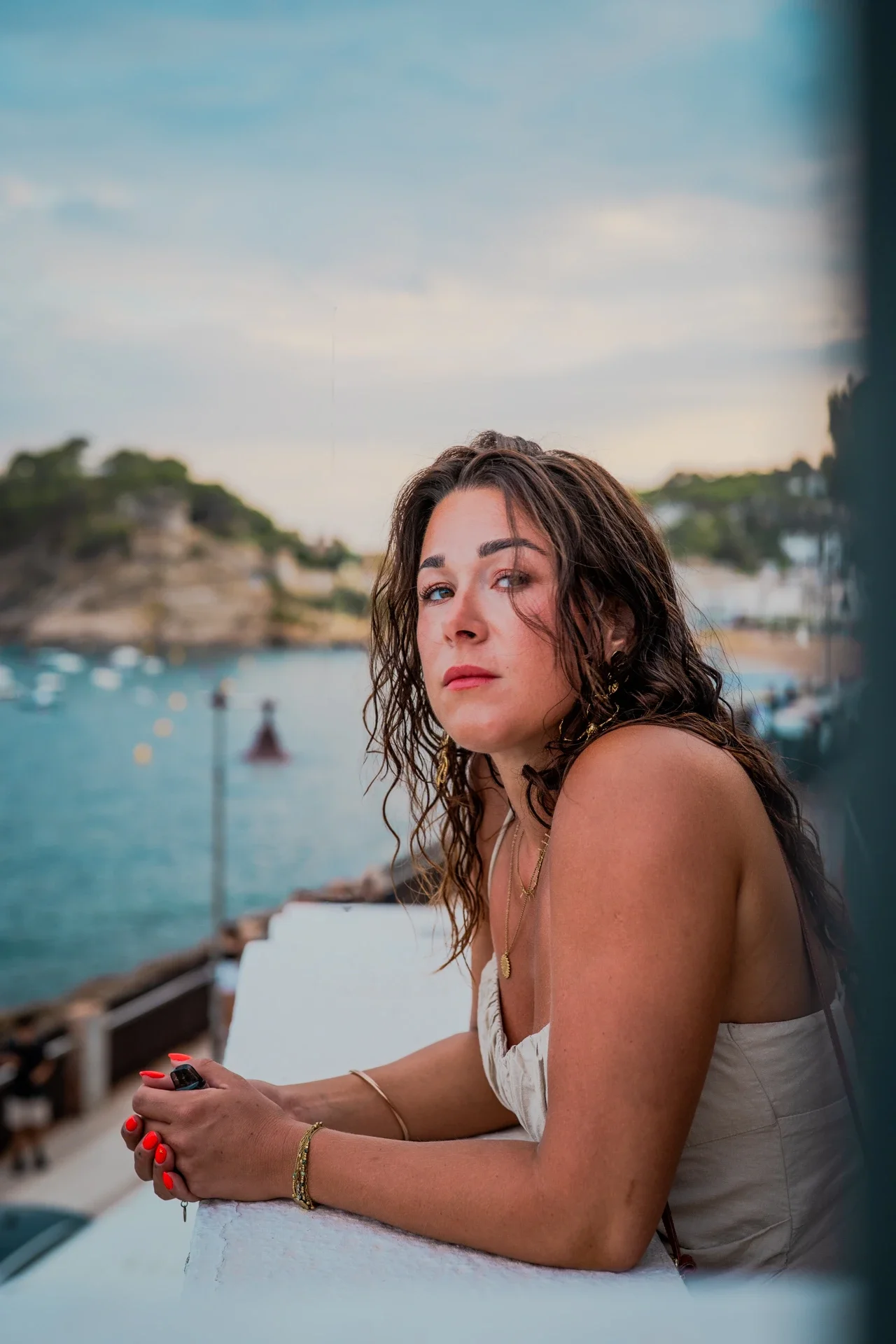 Une femme aux cheveux bouclés bruns regarde l'objectif, assise sur une terrasse avec vue sur la mer et la côte, au coucher du soleil.