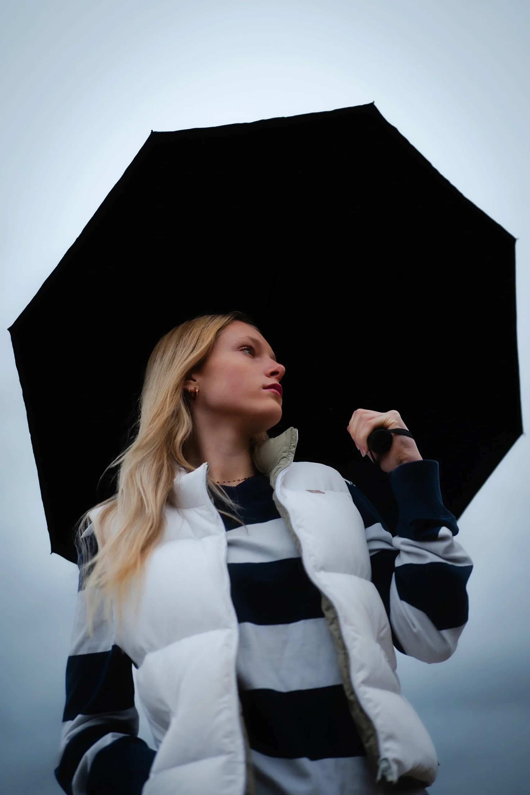 Jeune femme avec cheveux blonds portant un manteau blanc et un sweat à rayures noires et blanches, tenant un parapluie noir, regardant vers le haut, sur un ciel nuageux.