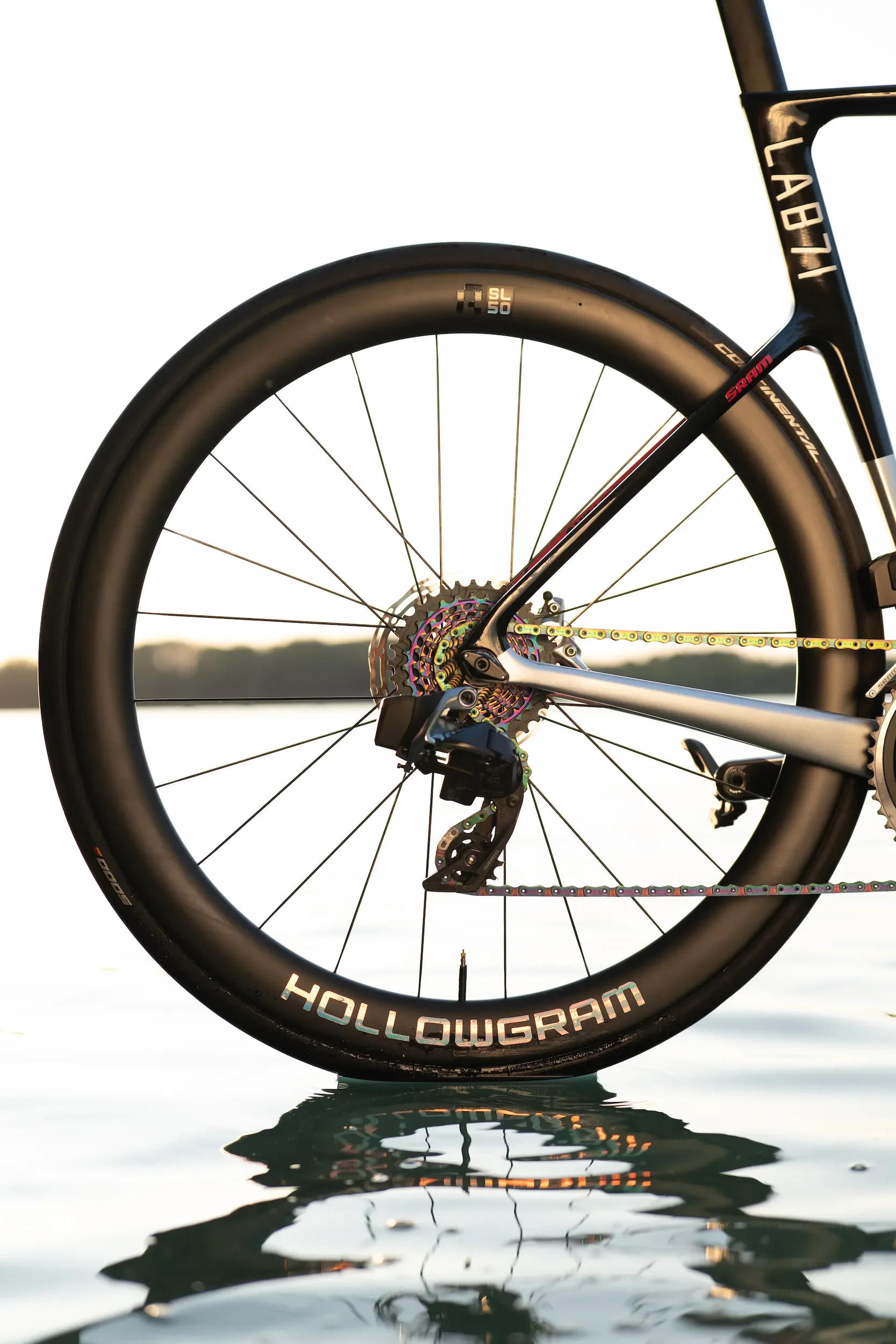 Vue partielle d'un vélo de course posé dans l'eau, avec le logo "HOLLOWGRAM" sur la roue et un paysage flou en arrière-plan.