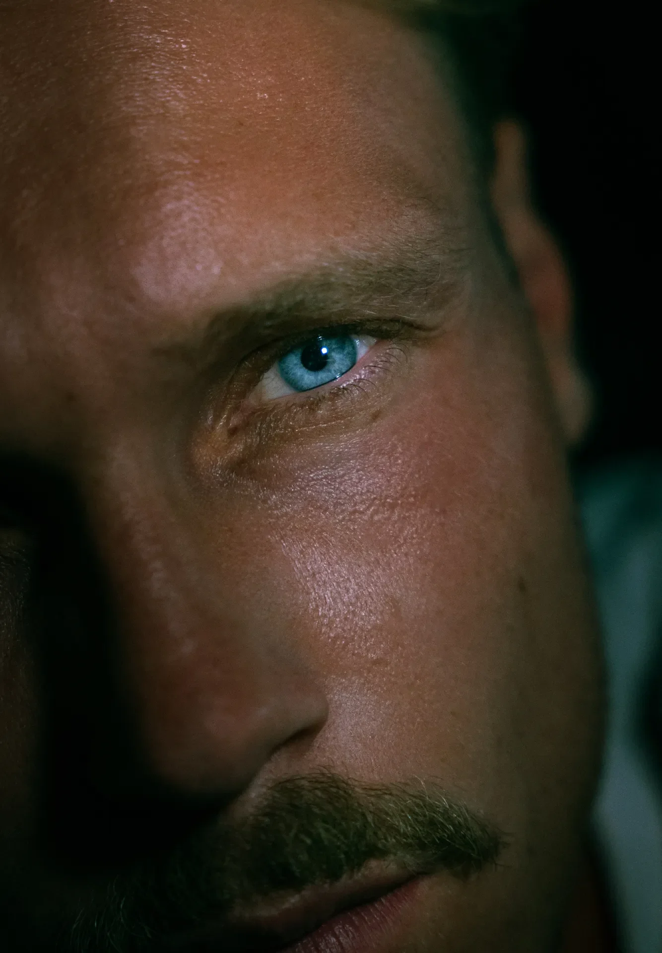 Zoom sur le visage d'un homme avec un œil bleu distinctif, peau bronzée et barbe, dans un éclairage sombre.