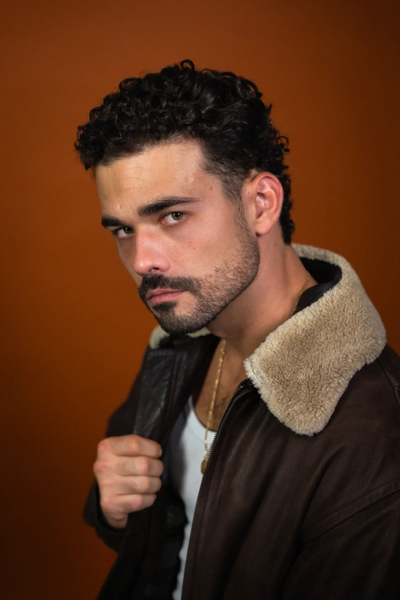 Un homme avec des cheveux bouclés, barbe et portant une veste en cuir avec un col en laine, pose contre un fond orange.