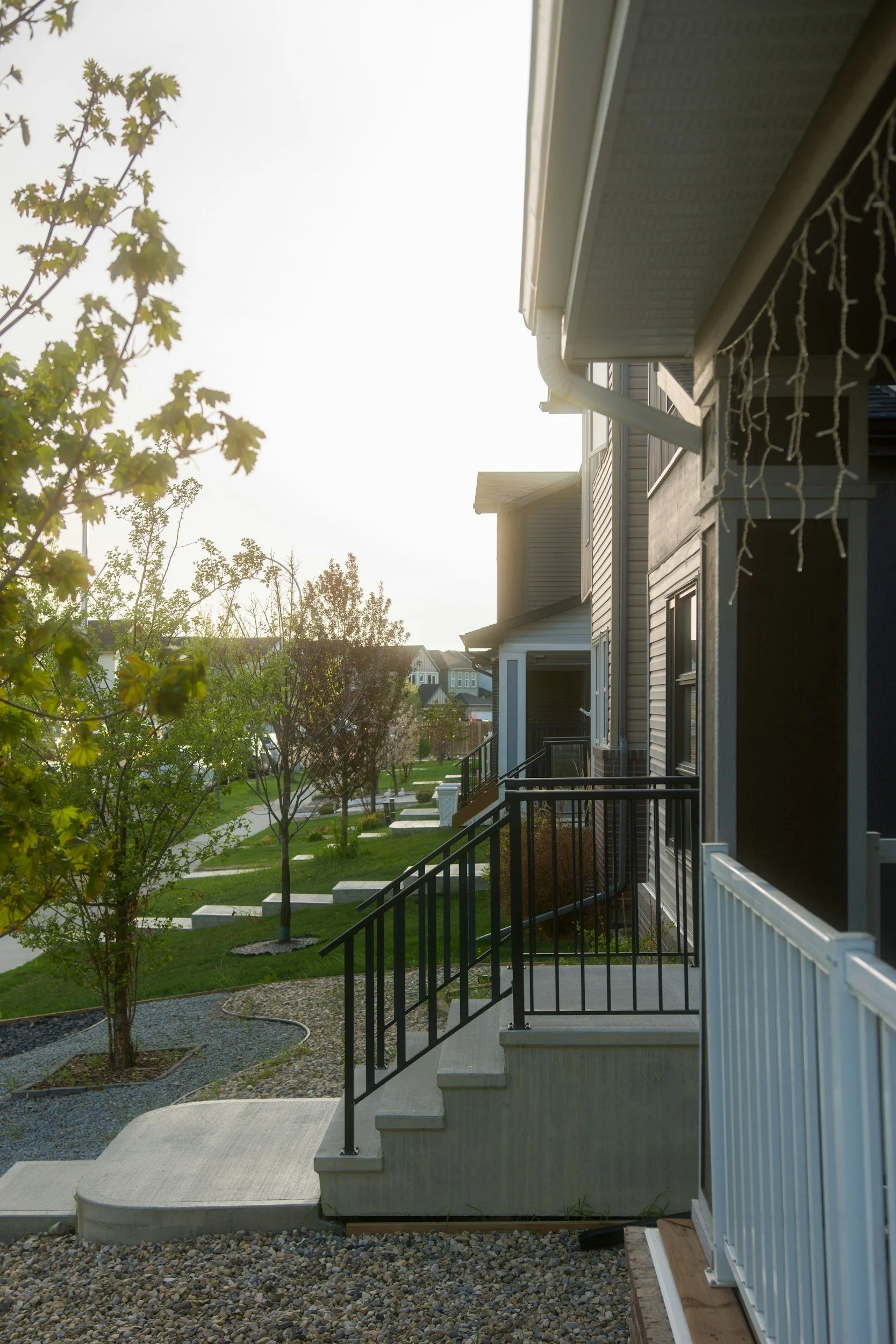 View of residential houses with front yard landscaping, trees, walkway, and stairway leading to a porch during daylight hours.