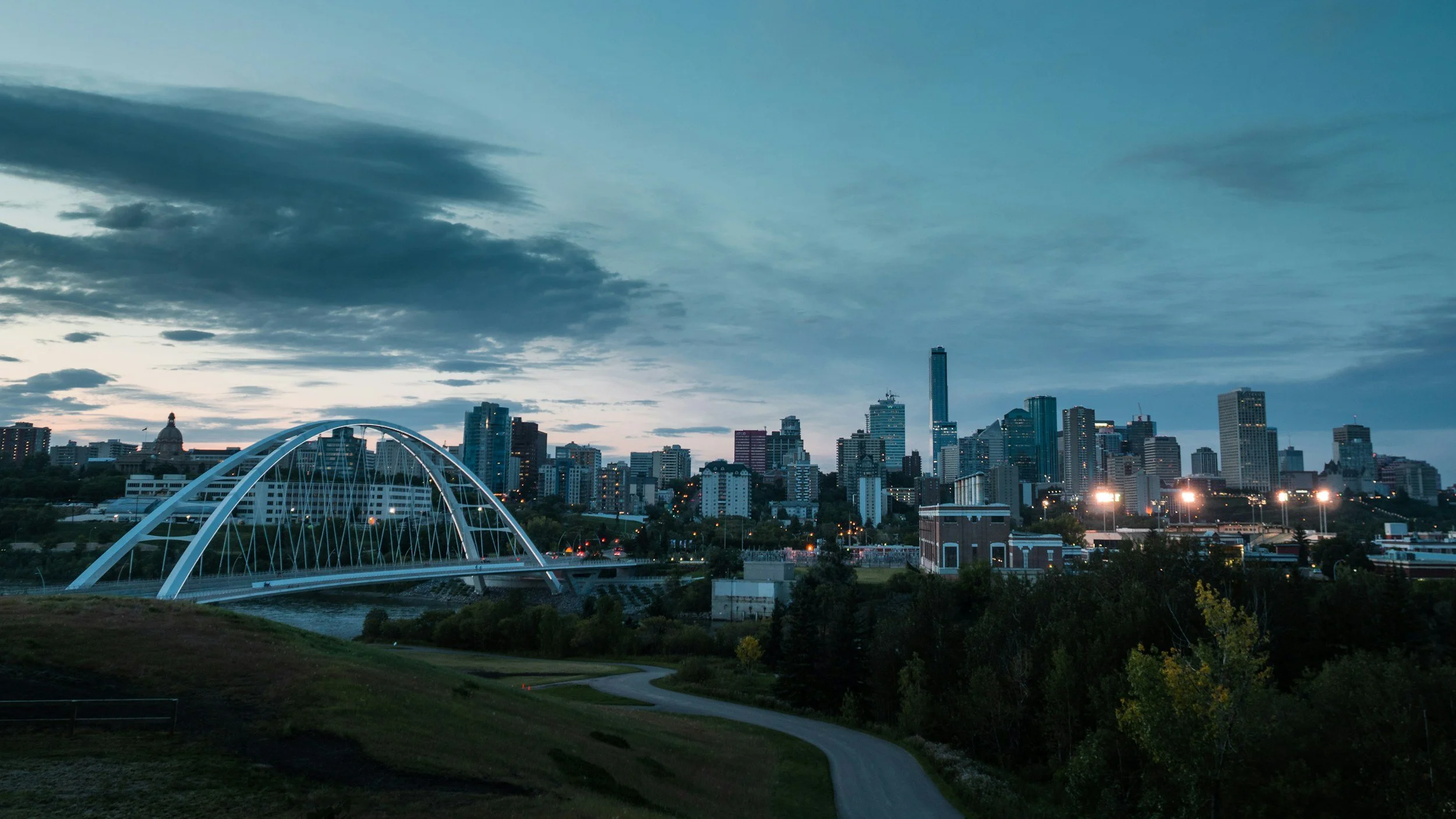 Skyline of a city at dusk with tall buildings, a bridge over a river, and a path in the foreground.