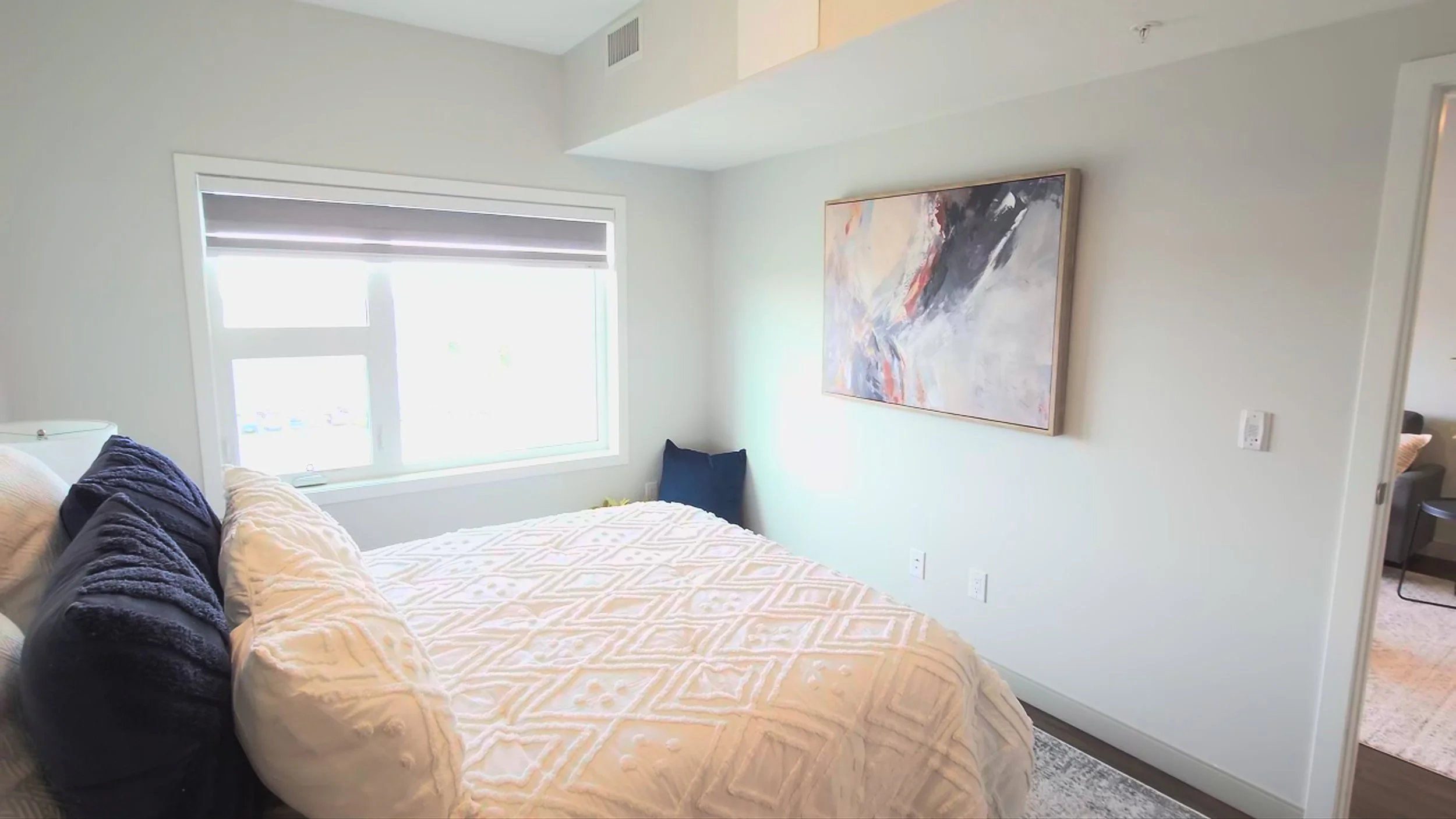 A bedroom with a bed, navy blue and white pillows, a large window, and an abstract painting on the wall.