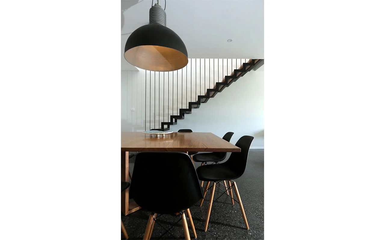 Modern dining area with a wooden table, black chairs, large industrial pendant light, and a floating staircase in the background.