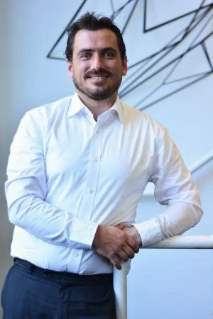Man in white shirt standing indoors, leaning on a railing, smiling