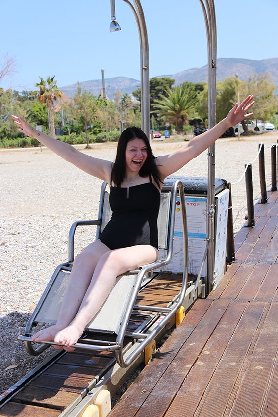 A woman in a black swimsuit sitting on a water slide with arms raised and a joyful expression, surrounded by a sunny outdoor park setting with trees and mountains in the background.