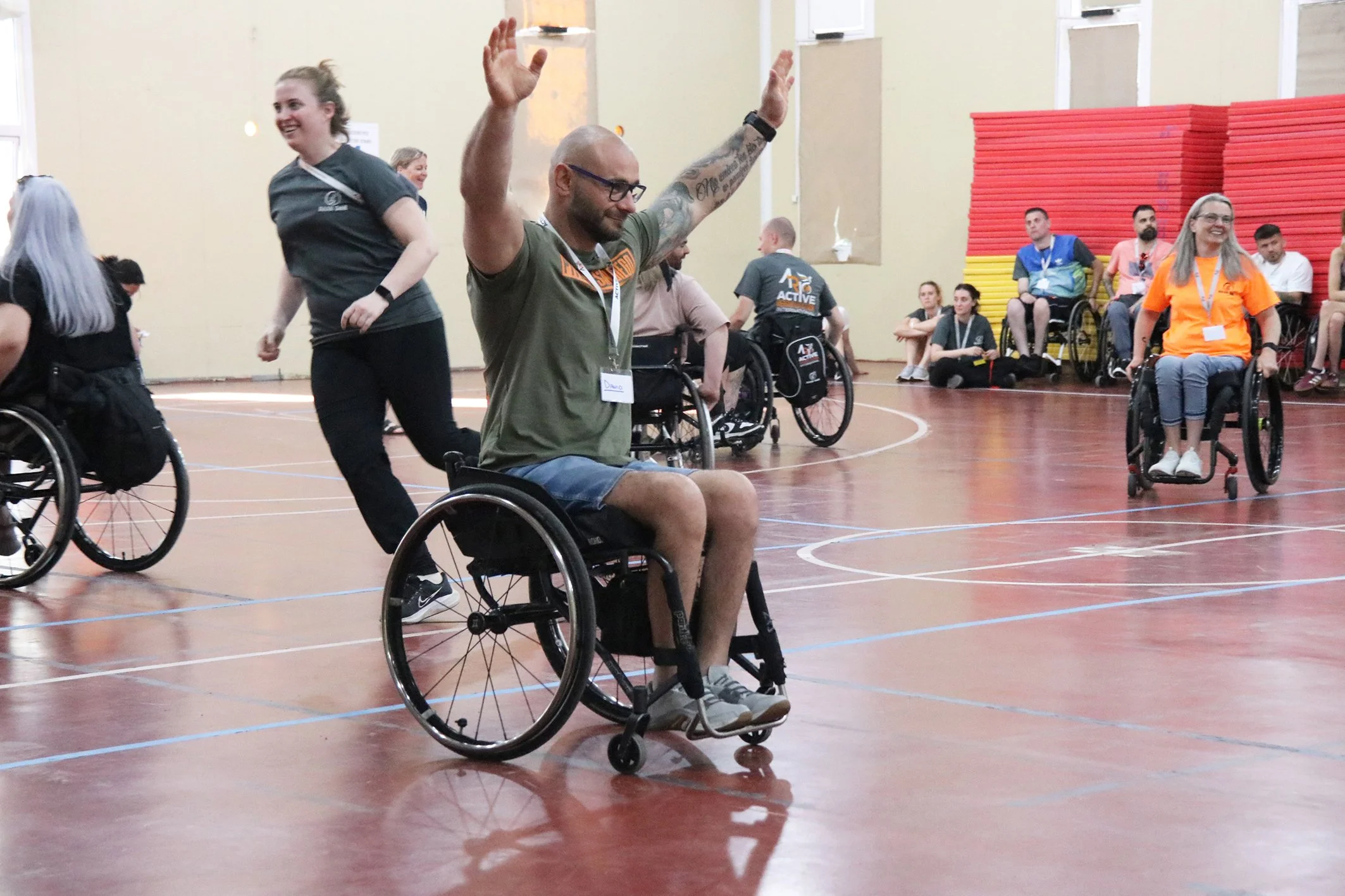 A man in a wheelchair with raised arms smiling during a wheelchair sports event, with other participants and spectators in the background.