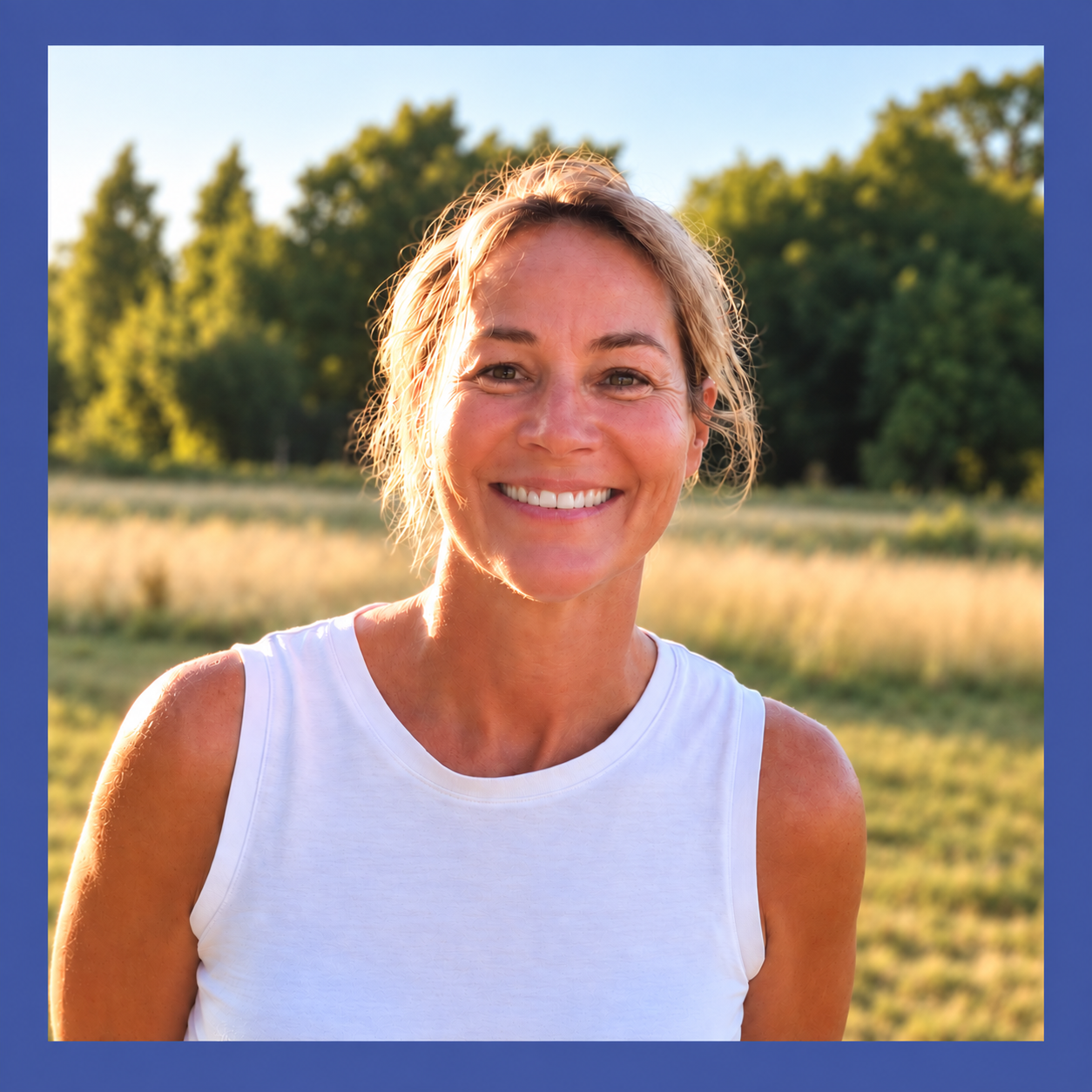 Smiling woman outdoors in a field with trees in the background during late afternoon sunshine.