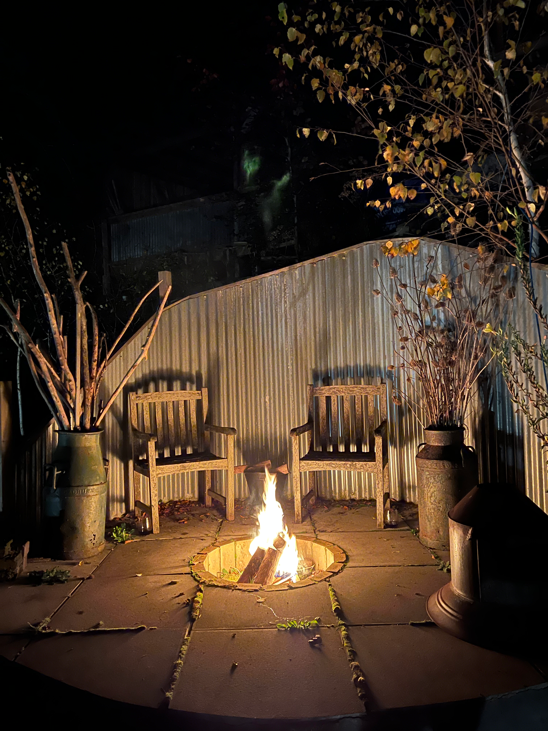 A backyard patio with a small fire in a circular fire pit. Two wooden chairs sit on either side of the fire, with tall potted plants on either side. A metal fence and trees with yellowing leaves are in the background, indicating an autumn setting at 