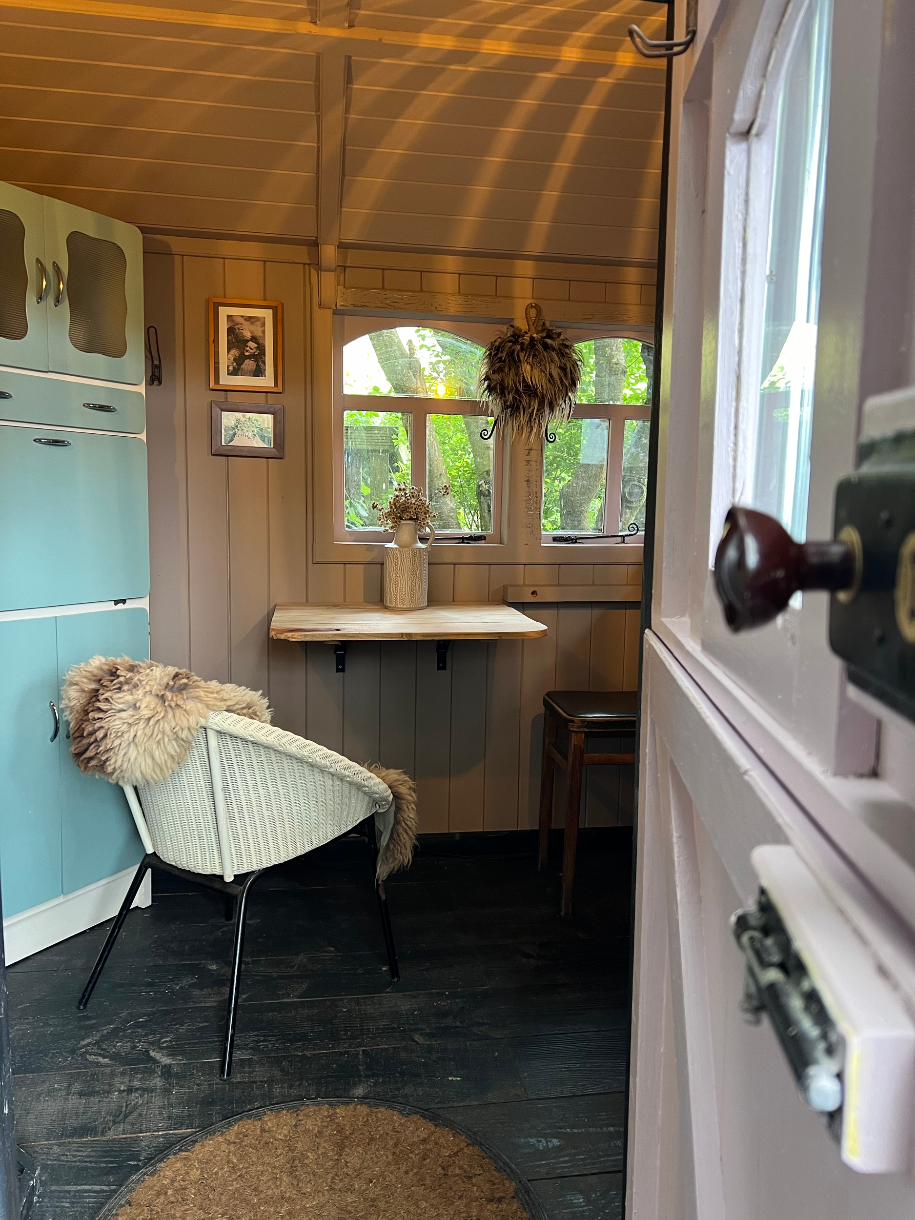 View of a cozy kitchen with a window overlooking a green outdoor scene, a white wicker chair with a faux fur throw, framed artwork on the wall, a wooden table under the window with a ceramic vase holding dried flowers, and a hanging feather decoratio