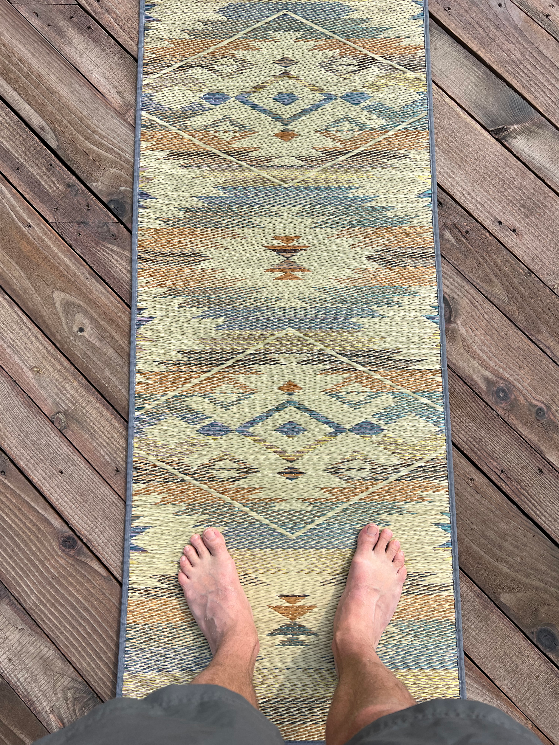 View of a person's bare feet standing on a colorful, patterned rug on a wooden deck floor, with the person's lower legs and shorts visible.