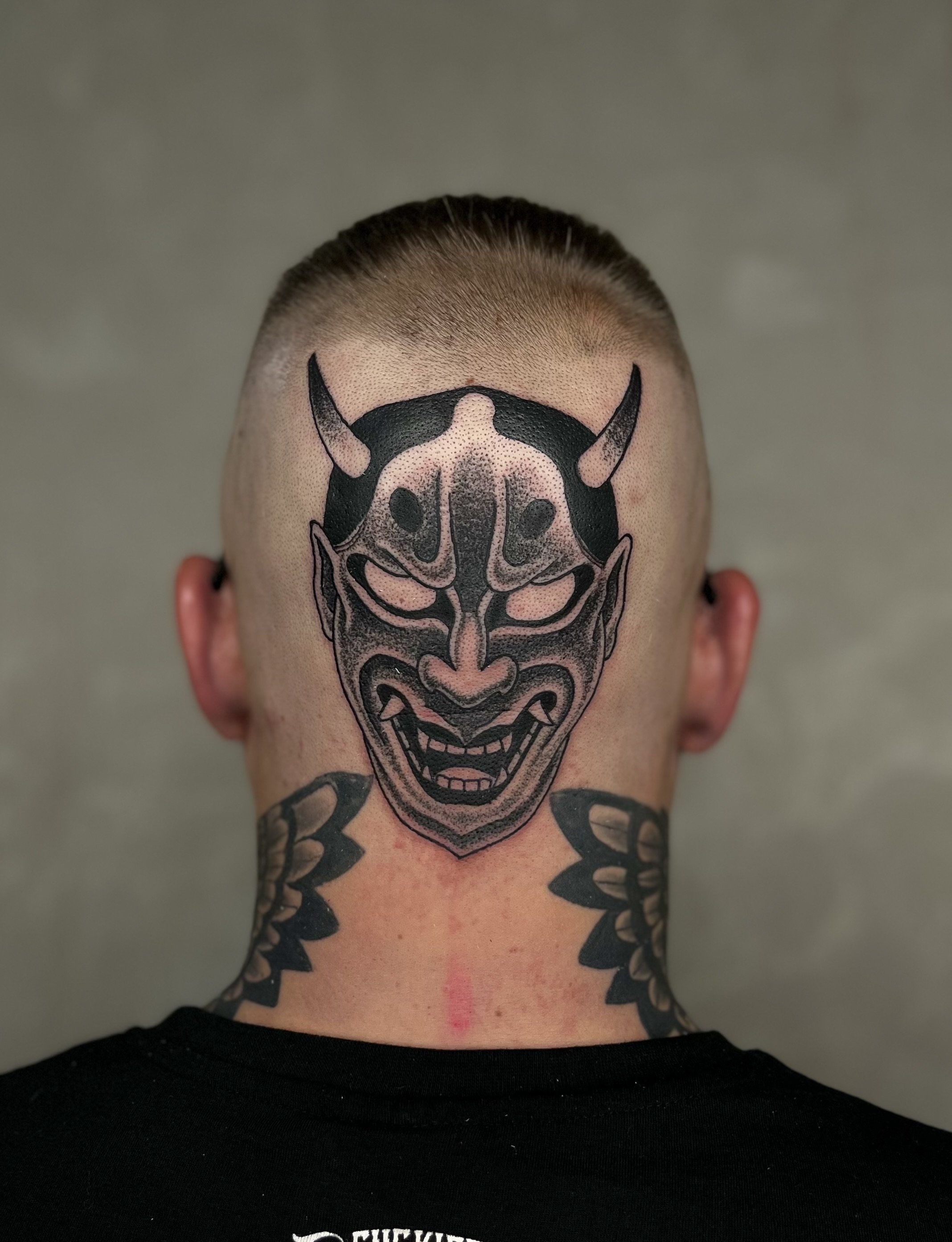 Tätowierung eines Oni-Dämonen auf dem Hinterkopf eines Mannes, mit scharfen Zähnen und Hörnern, darunter eine Tattoos-Verzierung am Nacken.