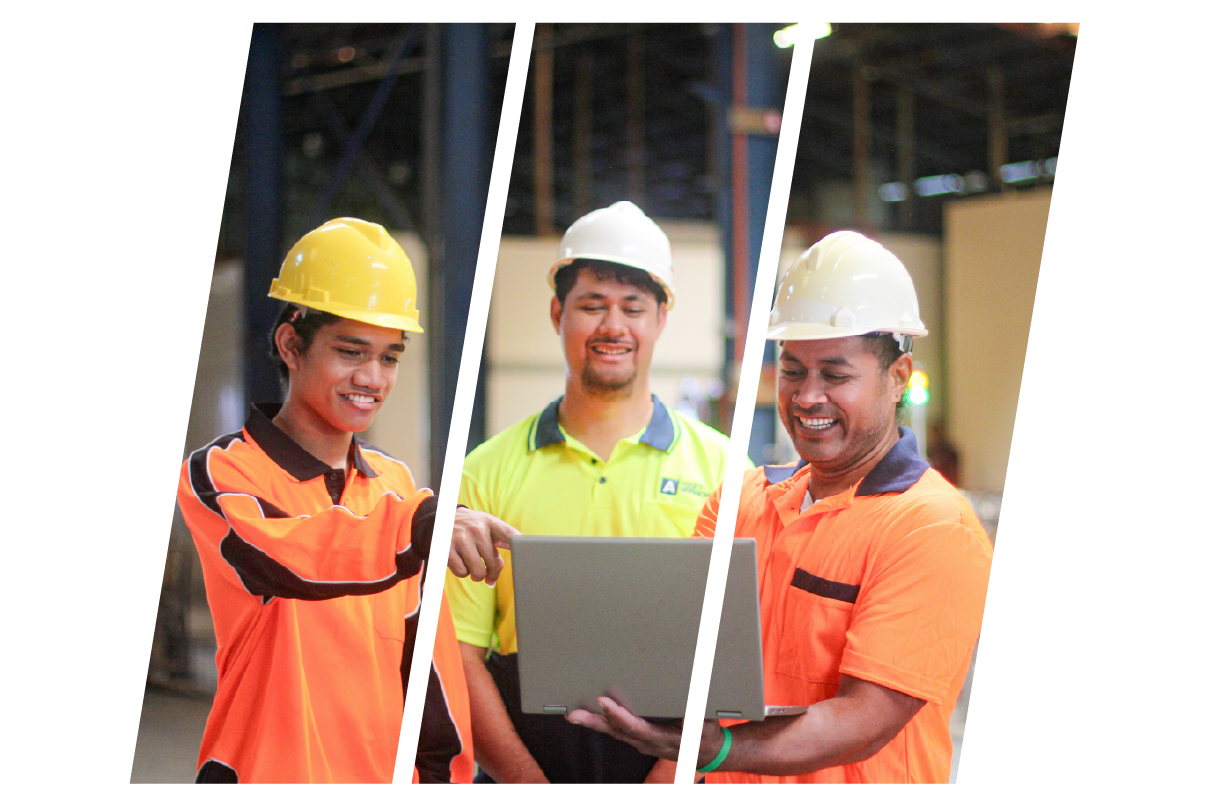 Three male workers in safety helmets and orange safety shirts standing in an industrial warehouse, looking at a gray laptop and smiling.