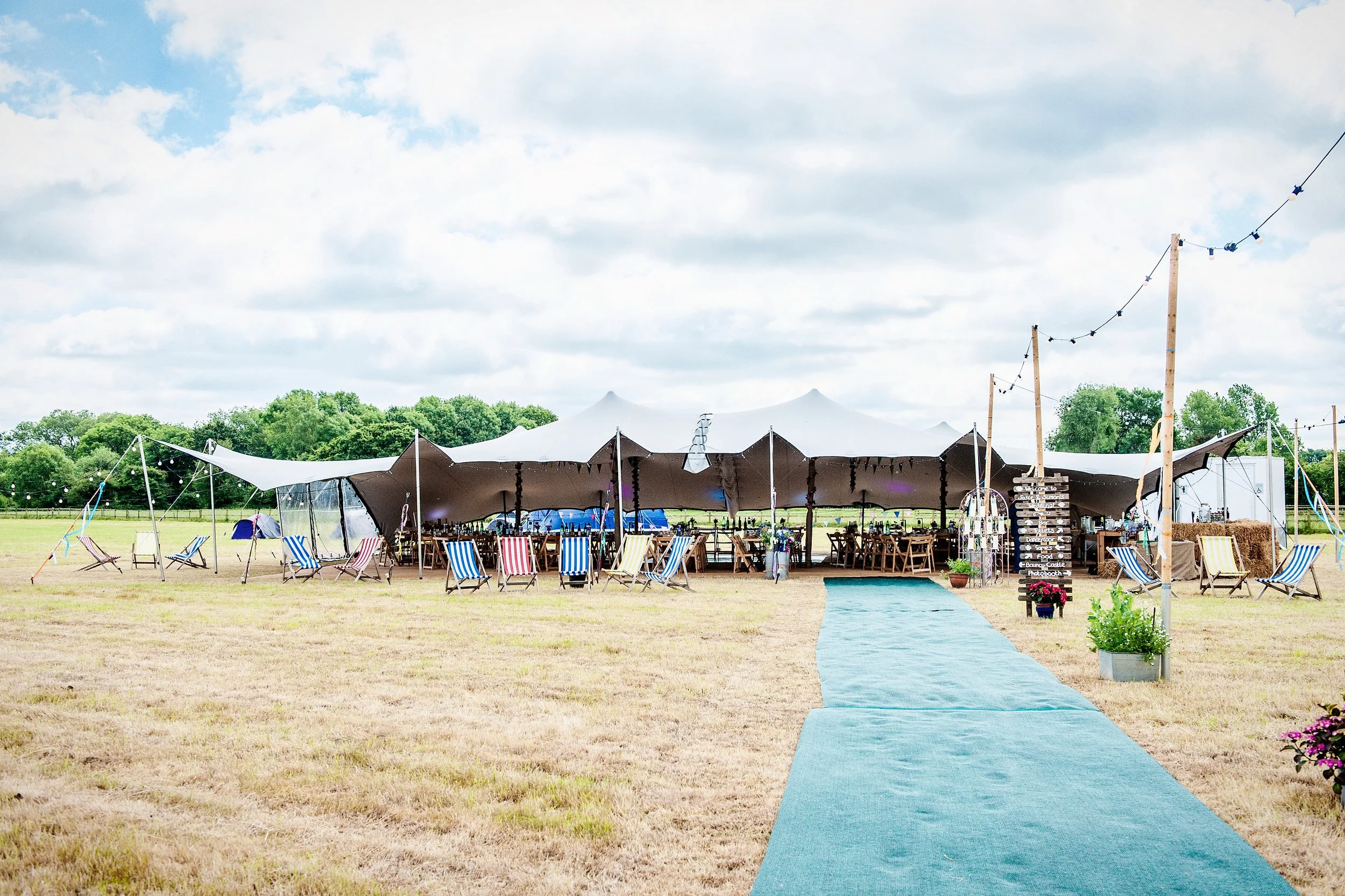 Outdoor wedding or event reception area under a large tent with blue and white chairs, a teal carpet walkway, string lights, and greenery in the background.