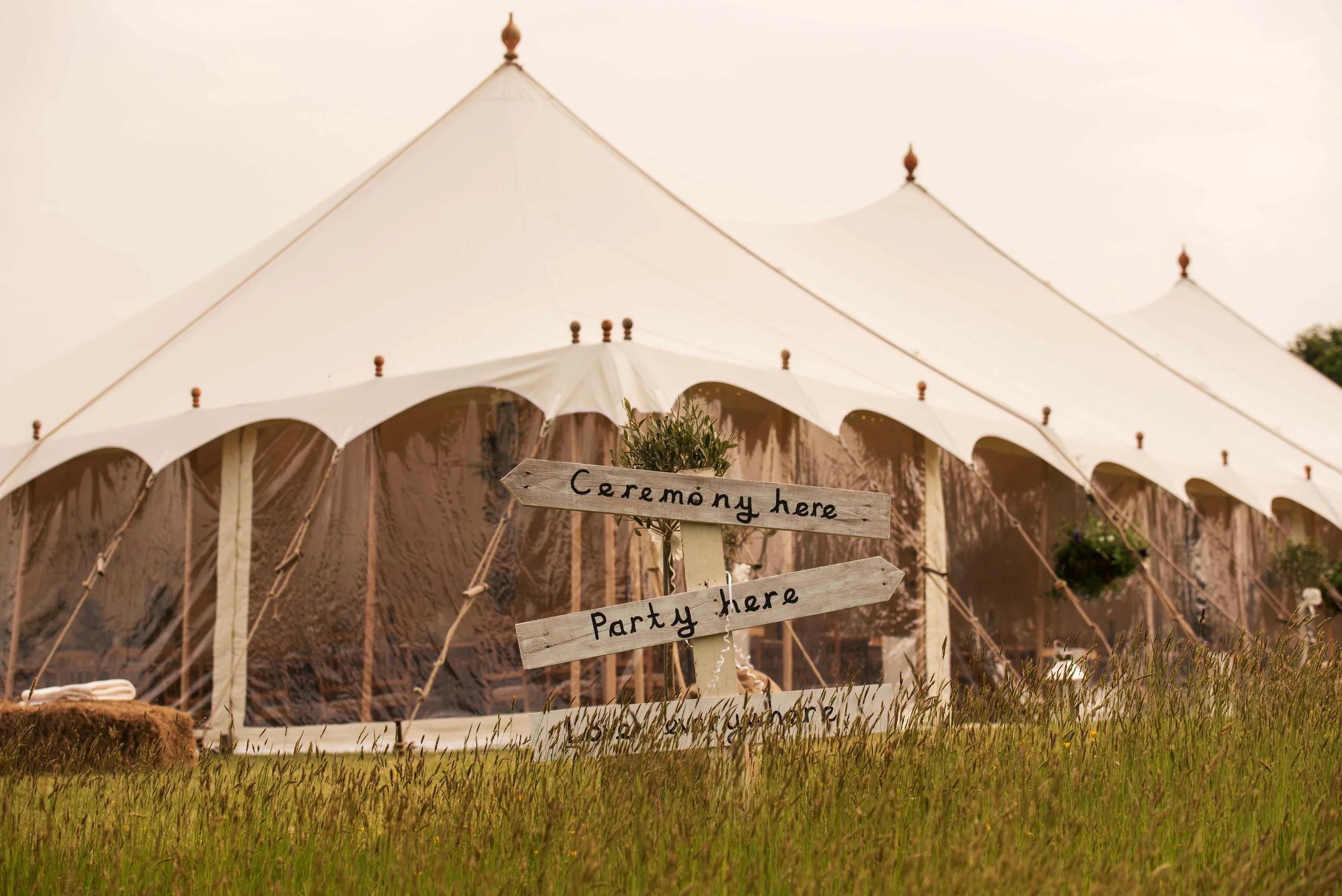 A large white event tent with pointed tops and wooden poles in an outdoor setting, surrounded by grass with a rustic wooden sign in front indicating directions for the ceremony and party areas.