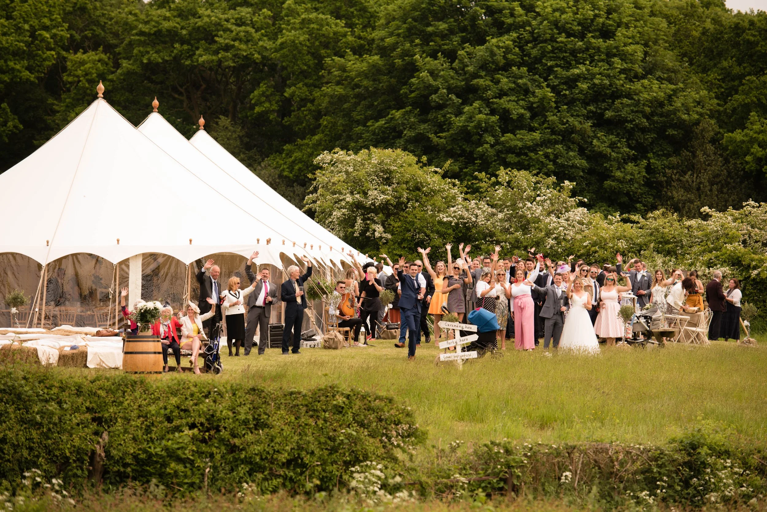 Group of wedding guests celebrating outdoors near a large white tent, with trees and greenery in the background.