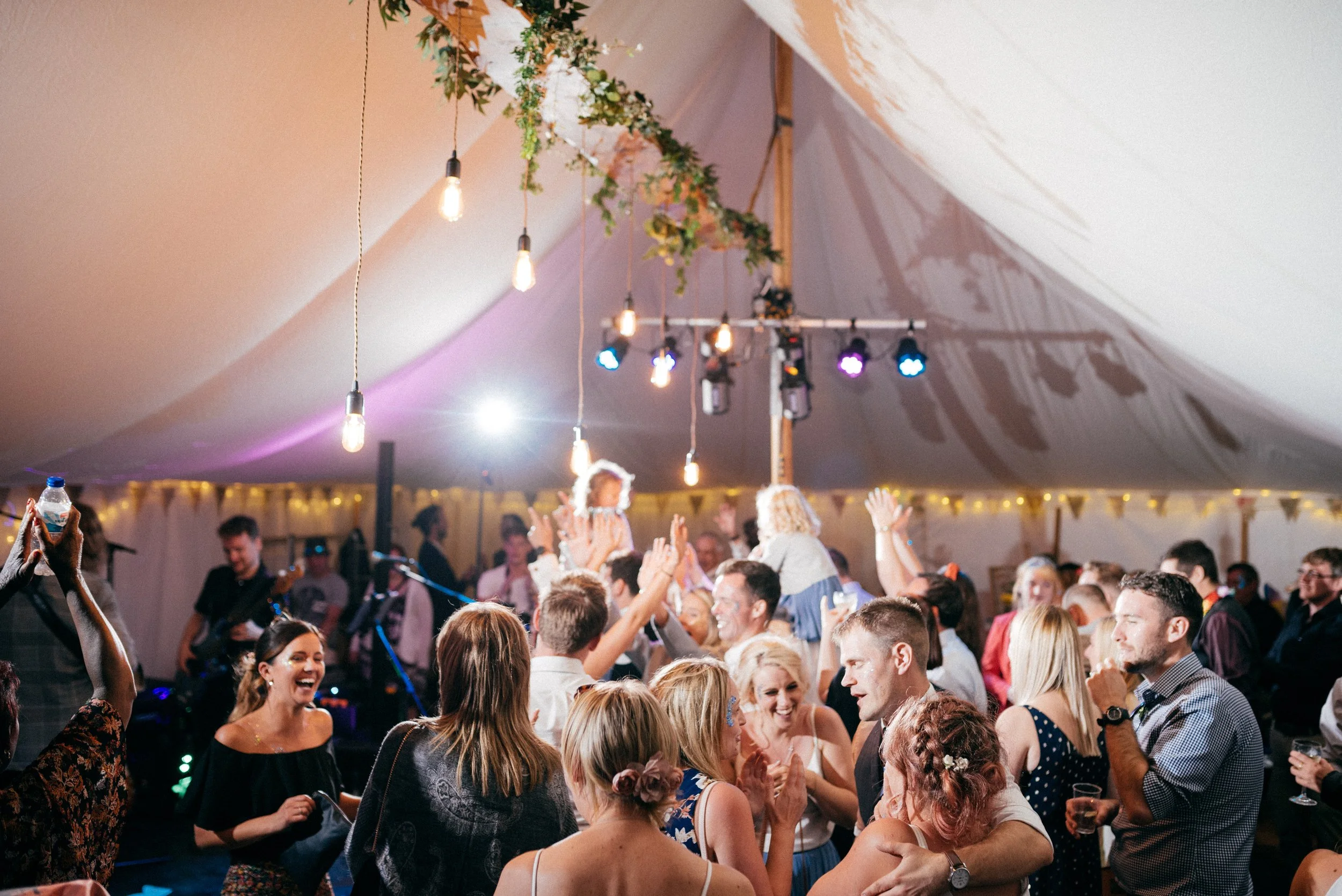 Guests dancing and celebrating at a lively party inside a decorated tent, with hanging lights, flowers, and a DJ playing music.