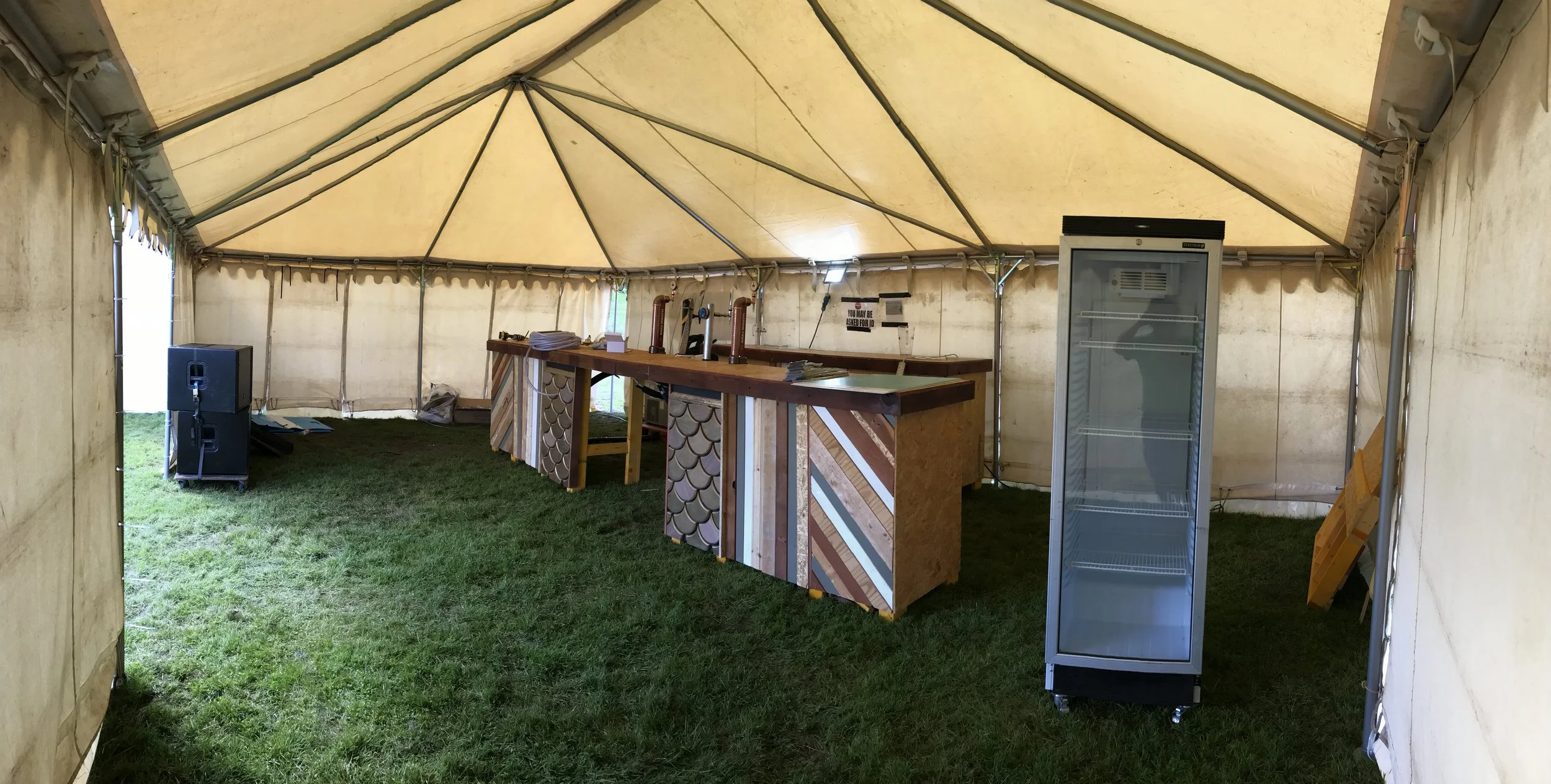 Interior of a beige event tent with a bar area, a large glass door refrigerator, and a speaker, set on grass.