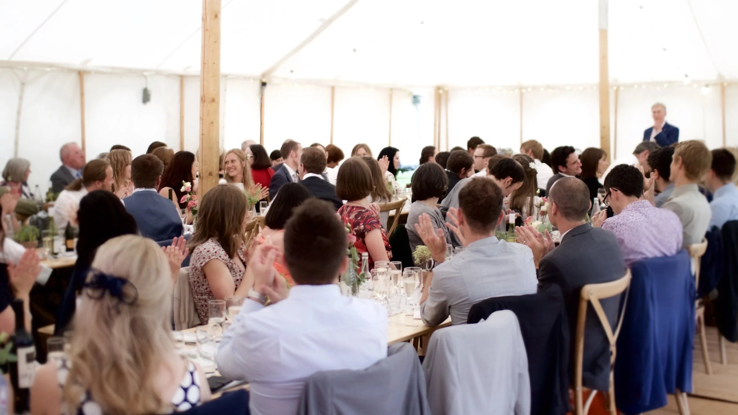 People attending a wedding reception inside a large white tent, sitting at long tables, clapping and smiling, in formal attire.