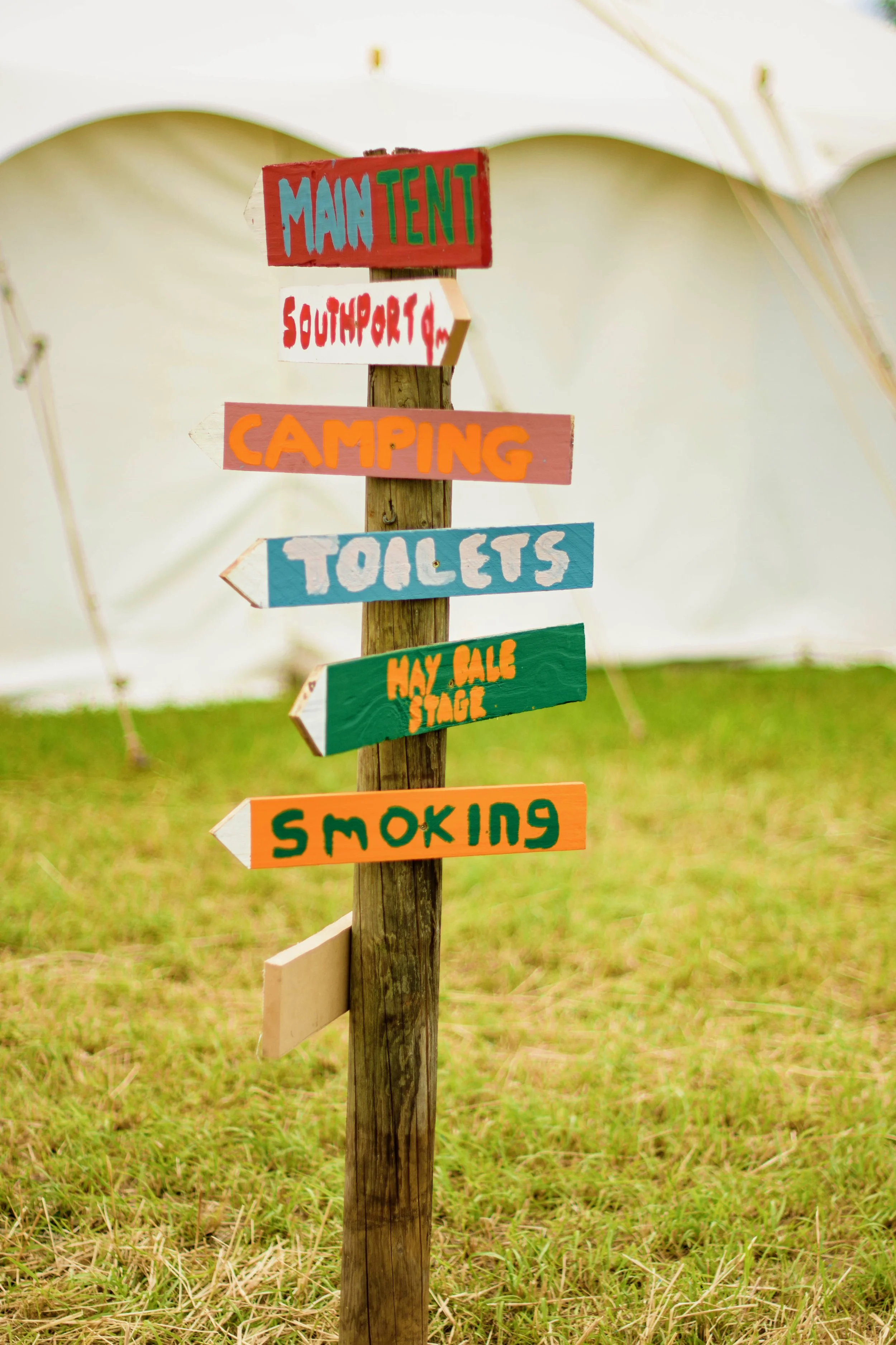 Colorful directional signpost with multiple signs pointing in different directions, including 'Main Tent,' 'South Port,' 'Camping,' 'Toilets,' 'Hay Bale Stage,' and 'Smoking,' set against a grassy outdoor background with tents.