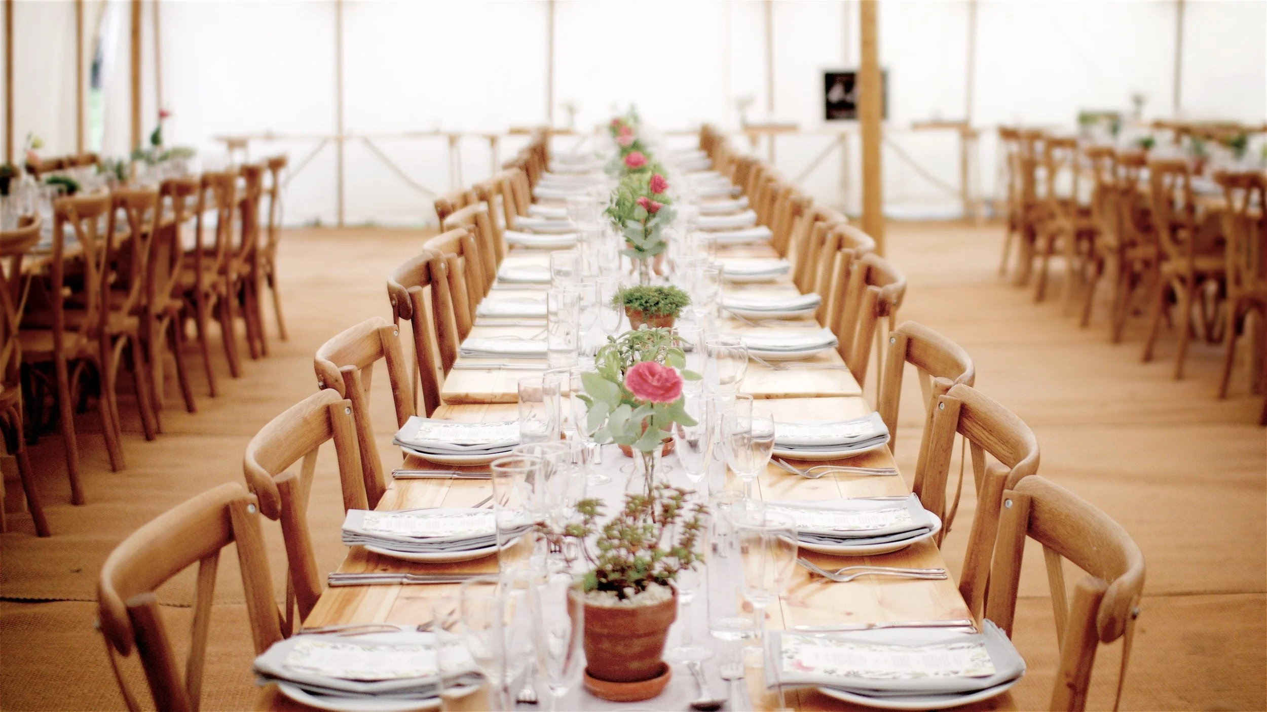 Long rectangular dining table set for a formal event with pink flowers and greenery centerpiece, surrounded by wooden chairs inside a white tent.
