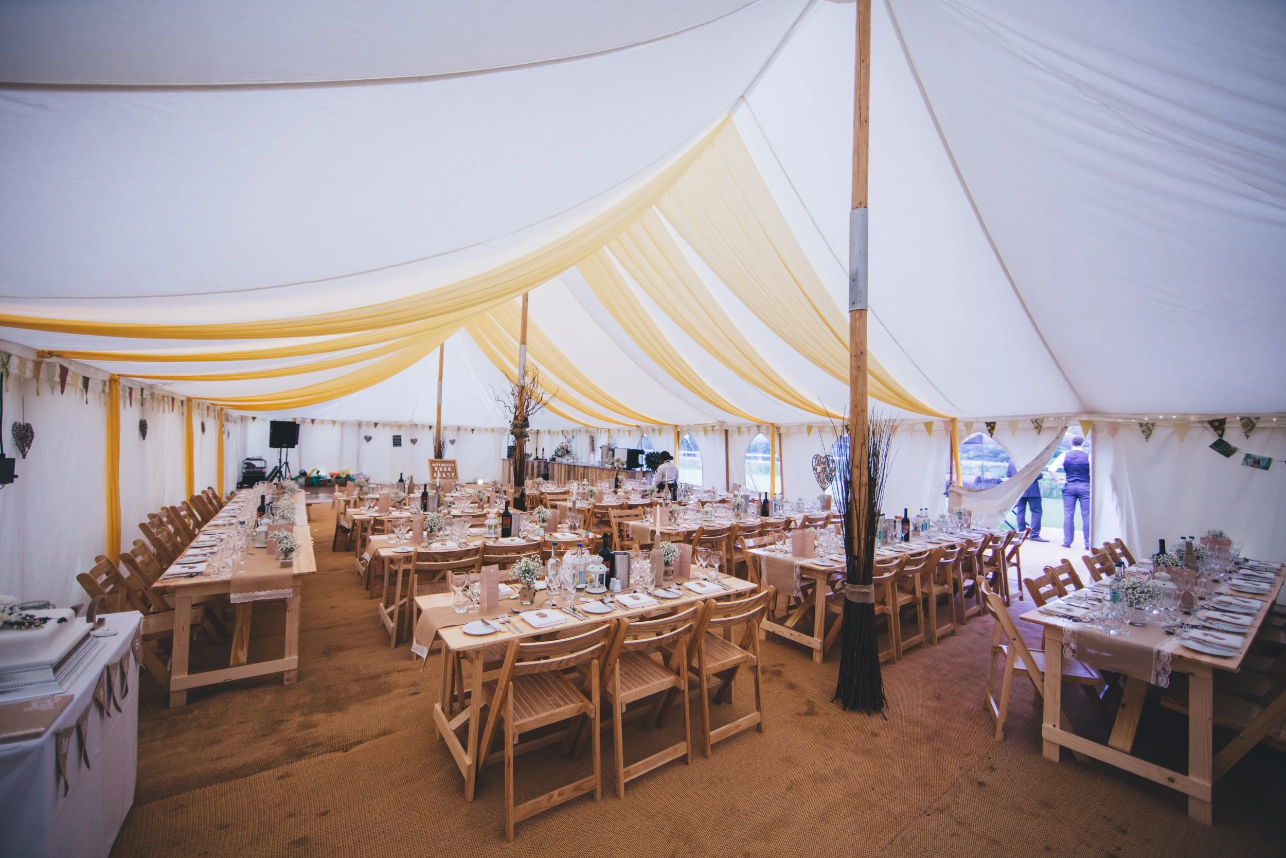 Large wedding reception tent decorated with yellow drapes, filled with long wooden tables and chairs, set with plates, glasses, and flowers, with some people standing outside near the entrance.