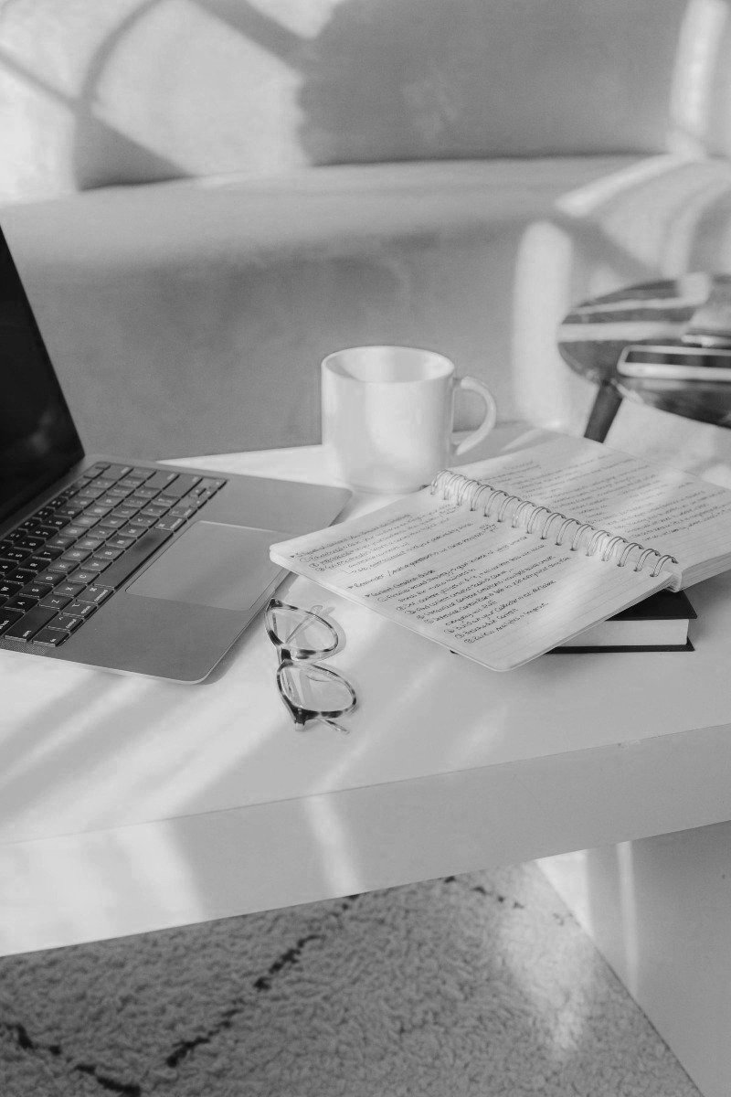 Black and white photo of a workspace with a laptop, an open notebook with handwritten notes, a pair of glasses, a ceramic mug, and a small desk lamp on a table.