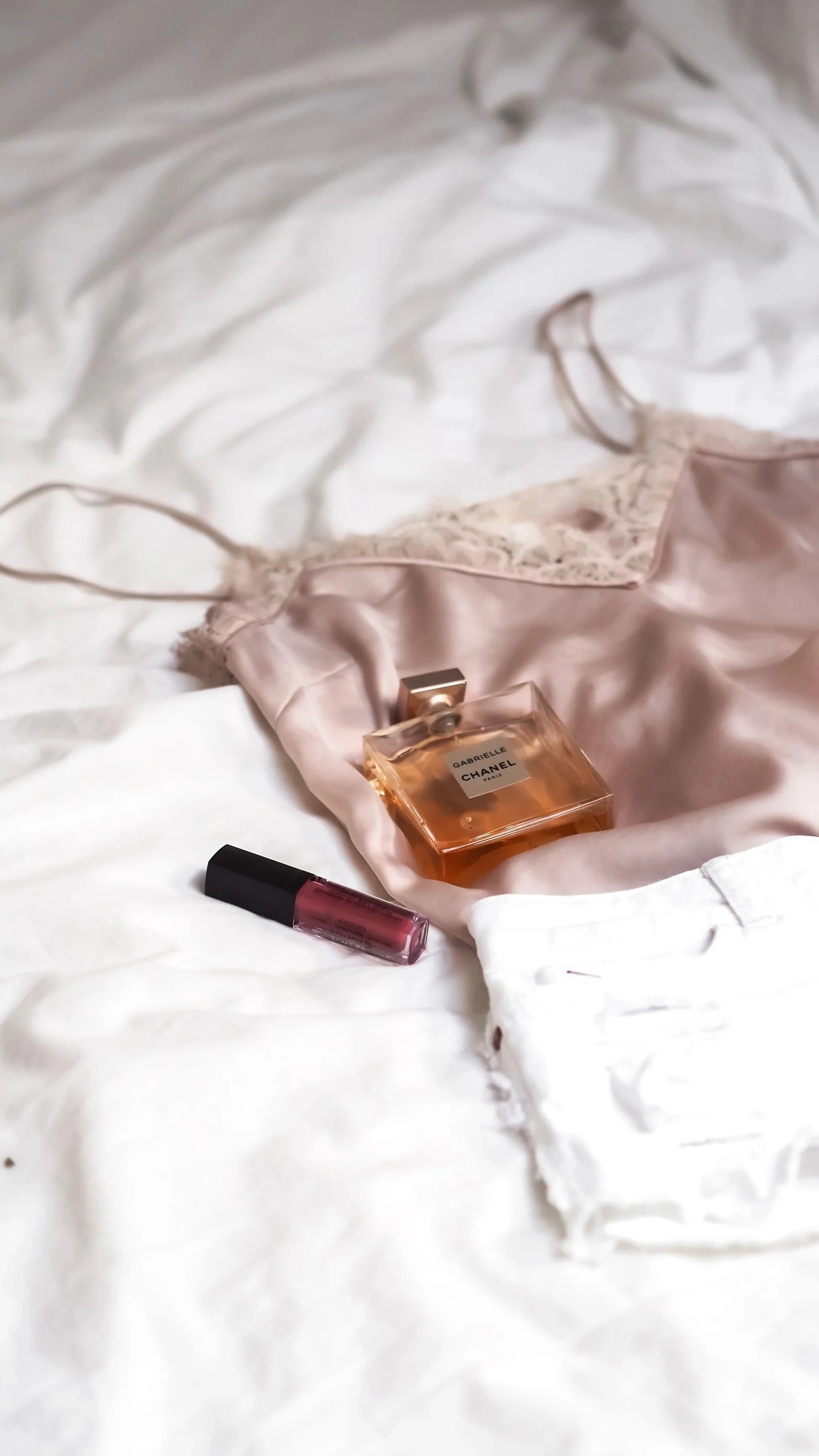 A pink silk camisole with lace trim, a bottle of Gabrielle Chanel perfume, and a small tube of lipstick placed on a white bedspread.