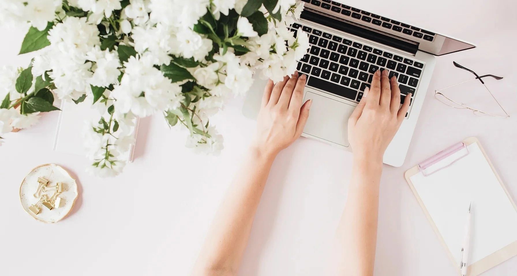 A person working on a laptop at a white desk, with a bouquet of white flowers, a pair of glasses, gold clips, and a notebook with a pen nearby.