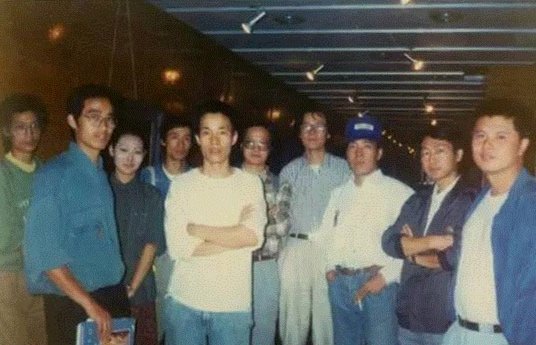 Group of people posing indoors, some with folded arms, wearing casual clothing, in a hallway with overhead lighting.