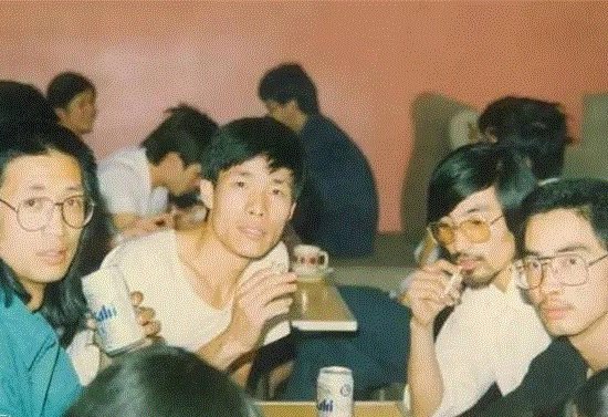 Group of people sitting at tables, some holding canned drinks, smoking in a casual indoor setting.