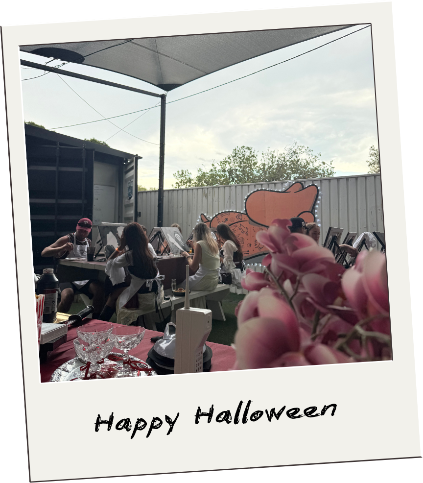 Outdoor Halloween celebration with people sitting at a long table, decorated with pink flowers and Halloween-themed decorations, under a shade sail on a cloudy day.