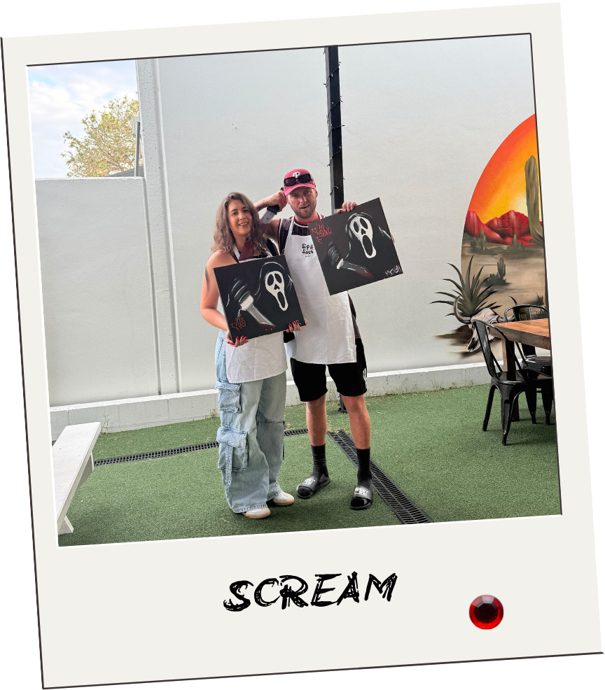 Two people, one woman and one man, standing outdoors holding horror movie-themed artwork with Ghostface mask. They are posing for a photo with a wall and desert landscape mural in the background.