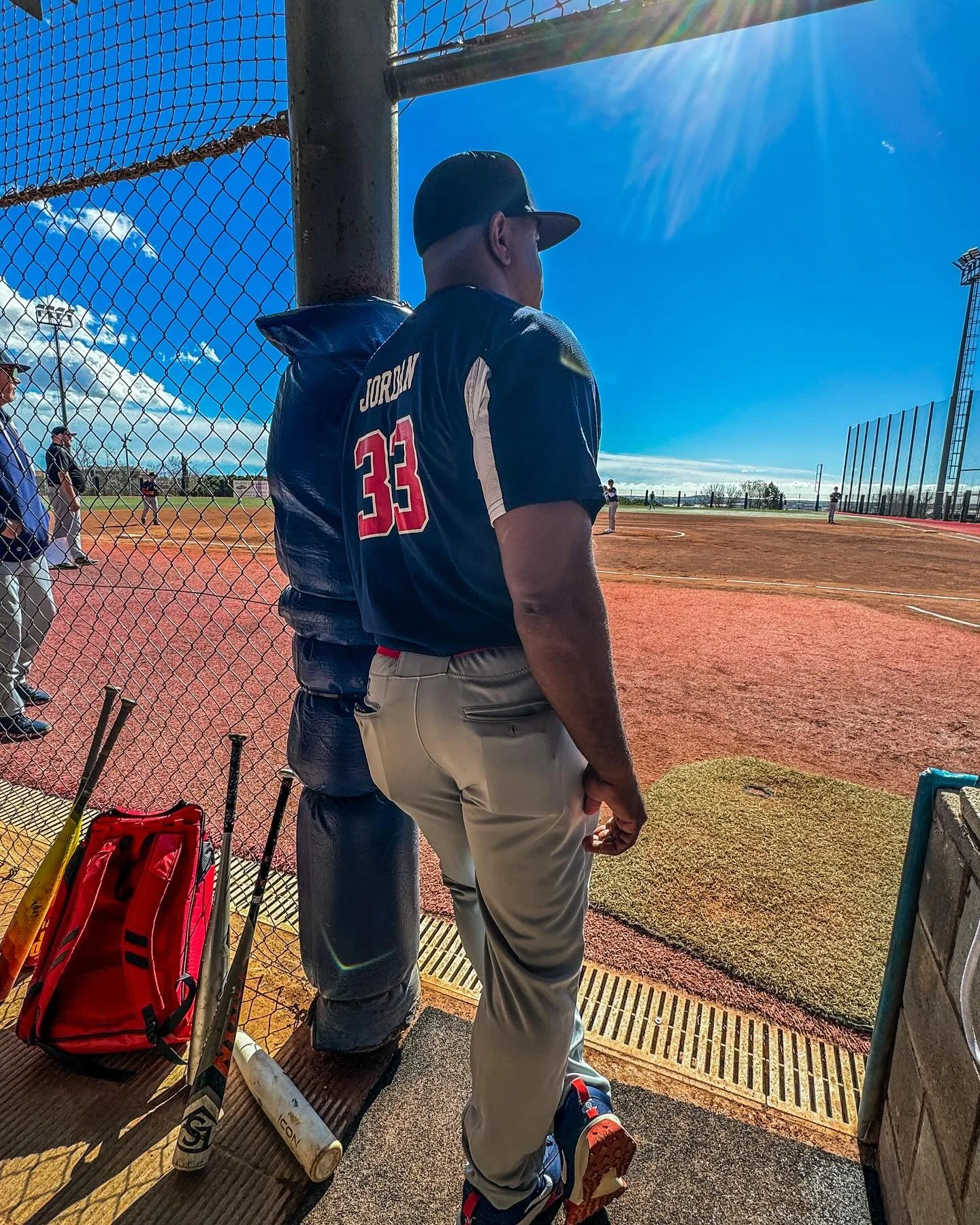 A baseball player wearing a dark jersey with the number 33 stands near a chain-link fence at a baseball field. Several bats and a red equipment bag are nearby. The sun shines brightly in a clear blue sky.