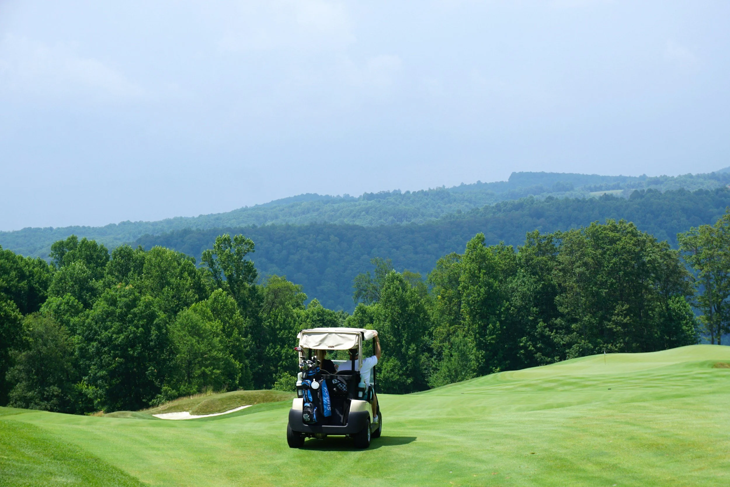 A golf course with a golf cart driving on the green, surrounded by trees and rolling hills in the distance.