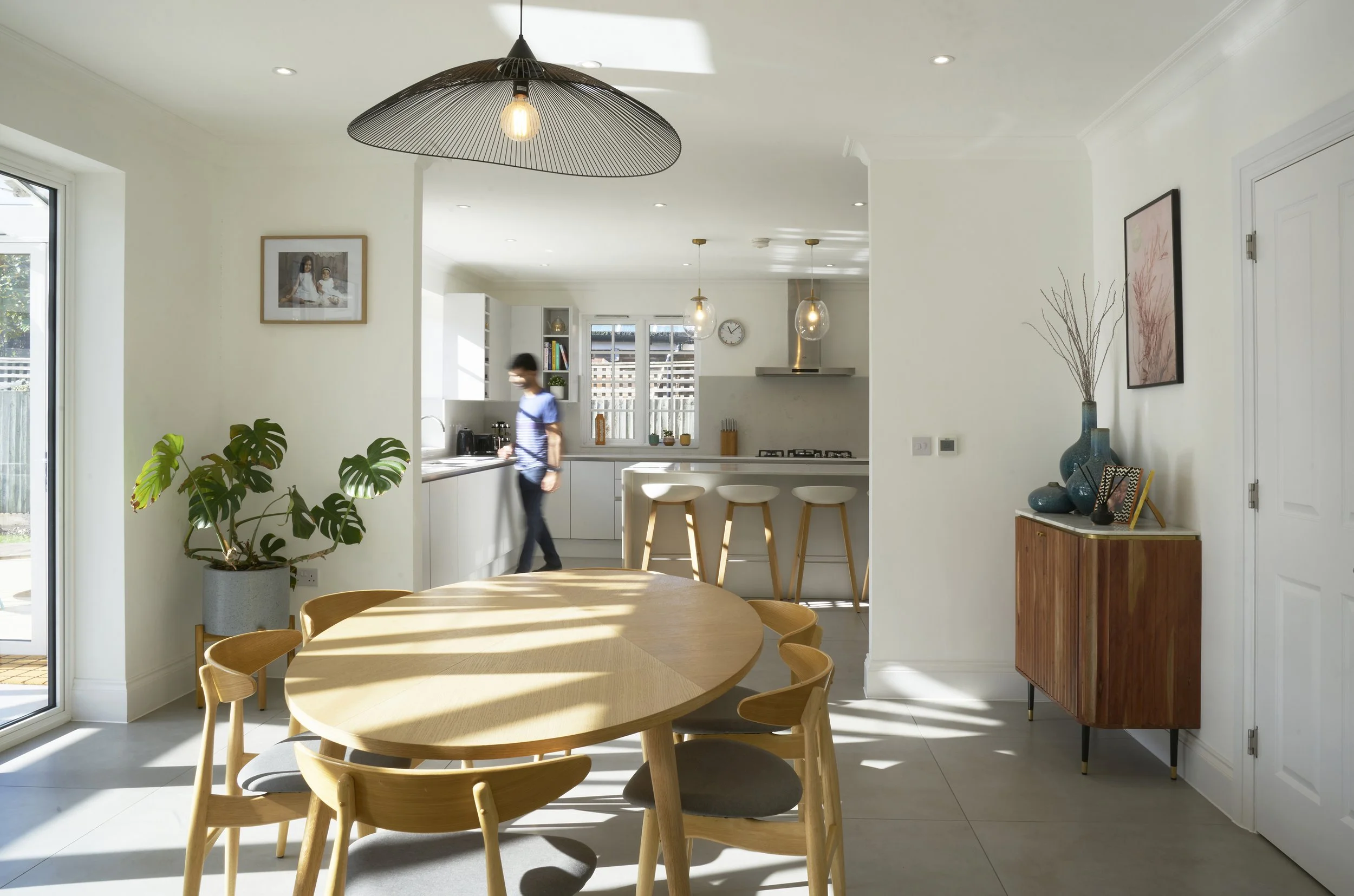 Interior view of a house with two young girls walking towards a door leading to a brightly lit room with a dining table and chairs, decorated with modern art and potted plants.