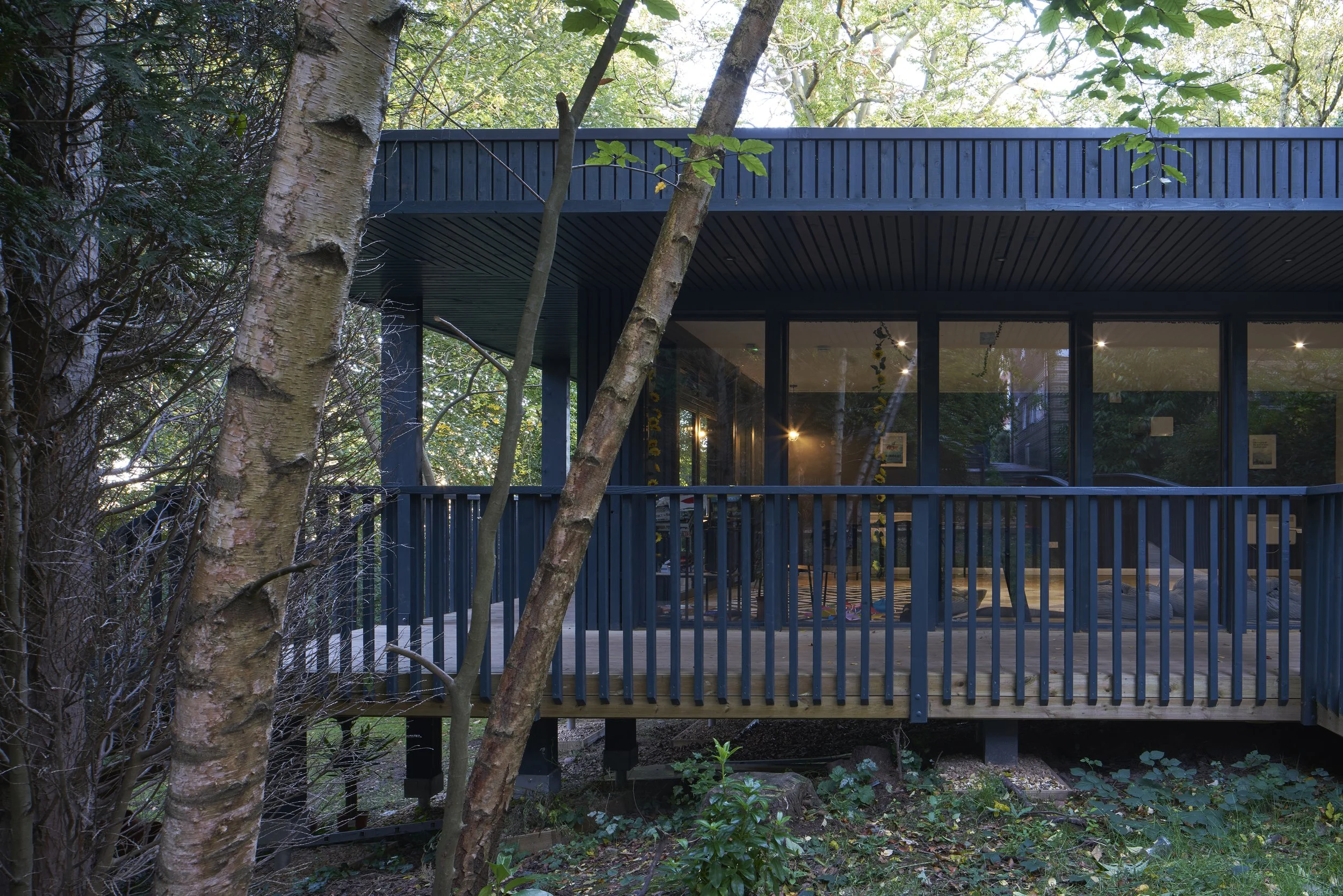 A modern house with large windows and a black exterior is surrounded by trees and bushes, with a deck and an interior view visible through the windows.