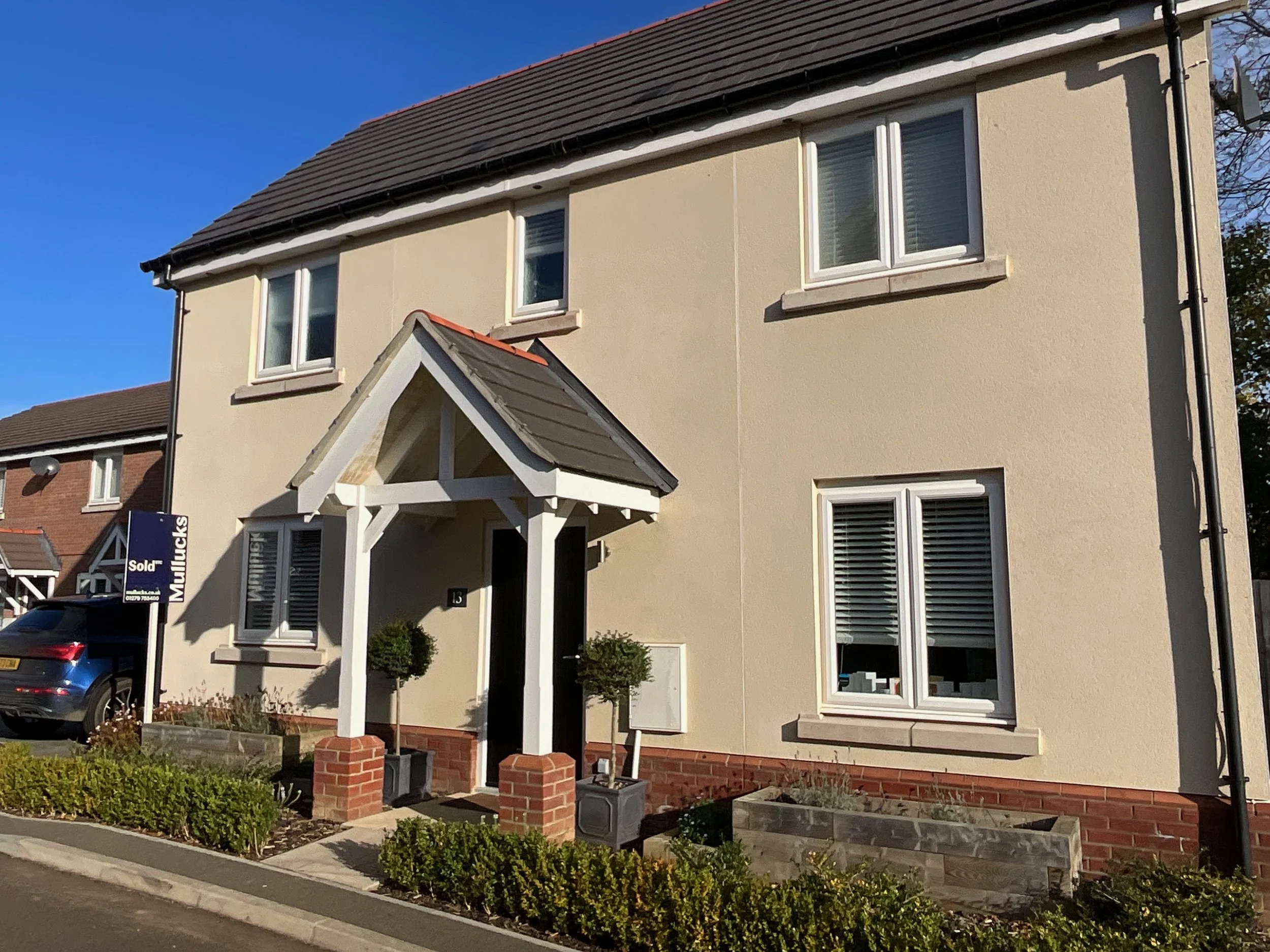 Two-story beige house with white window frames, small front porch with brick pillars, potted plants, and a sold sign in the yard