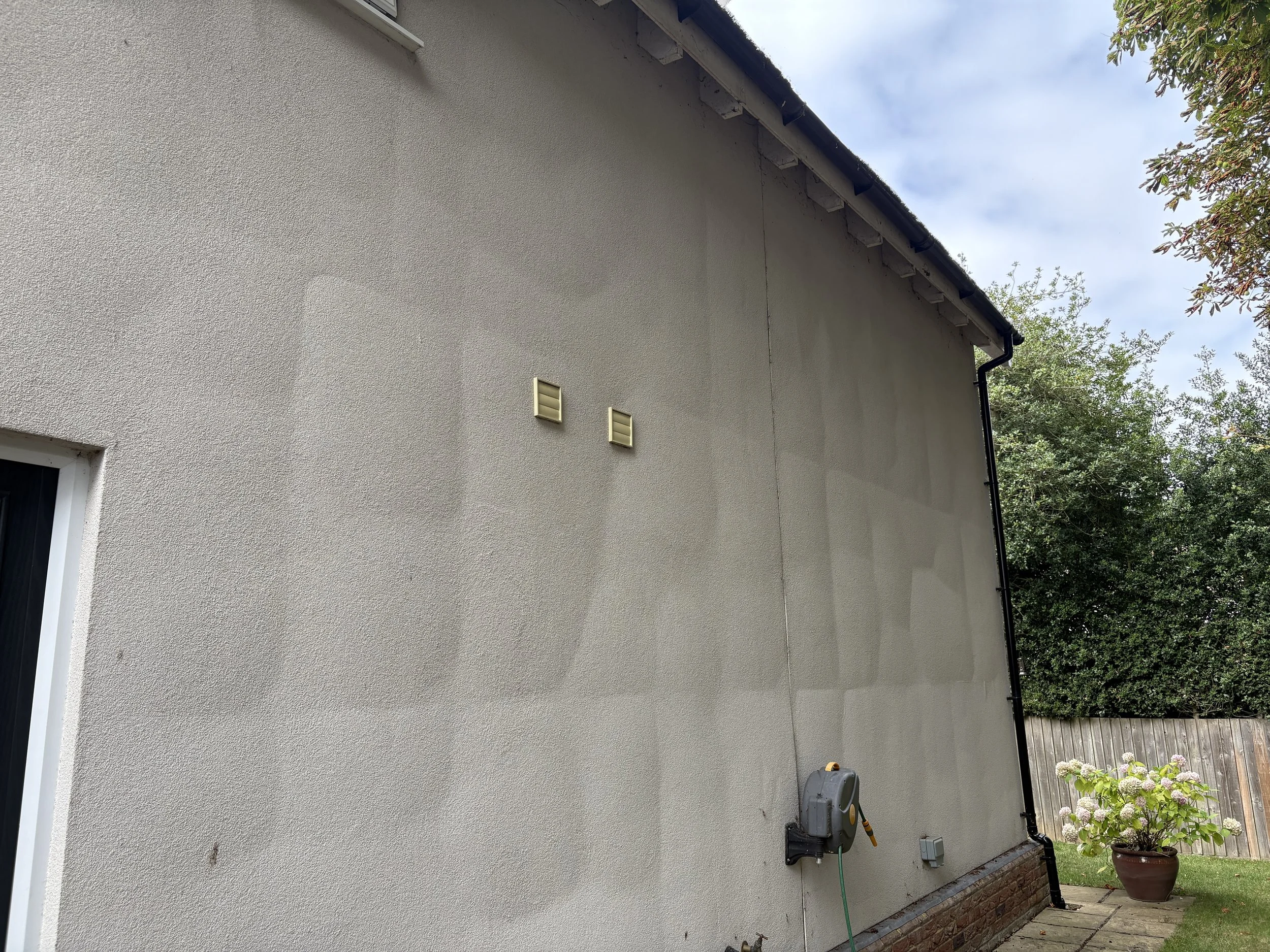 Exterior of a house with a stucco wall, two vent covers, a garden hose reel mounted on the wall, and a potted plant with flowers next to a wooden fence and green bushes.