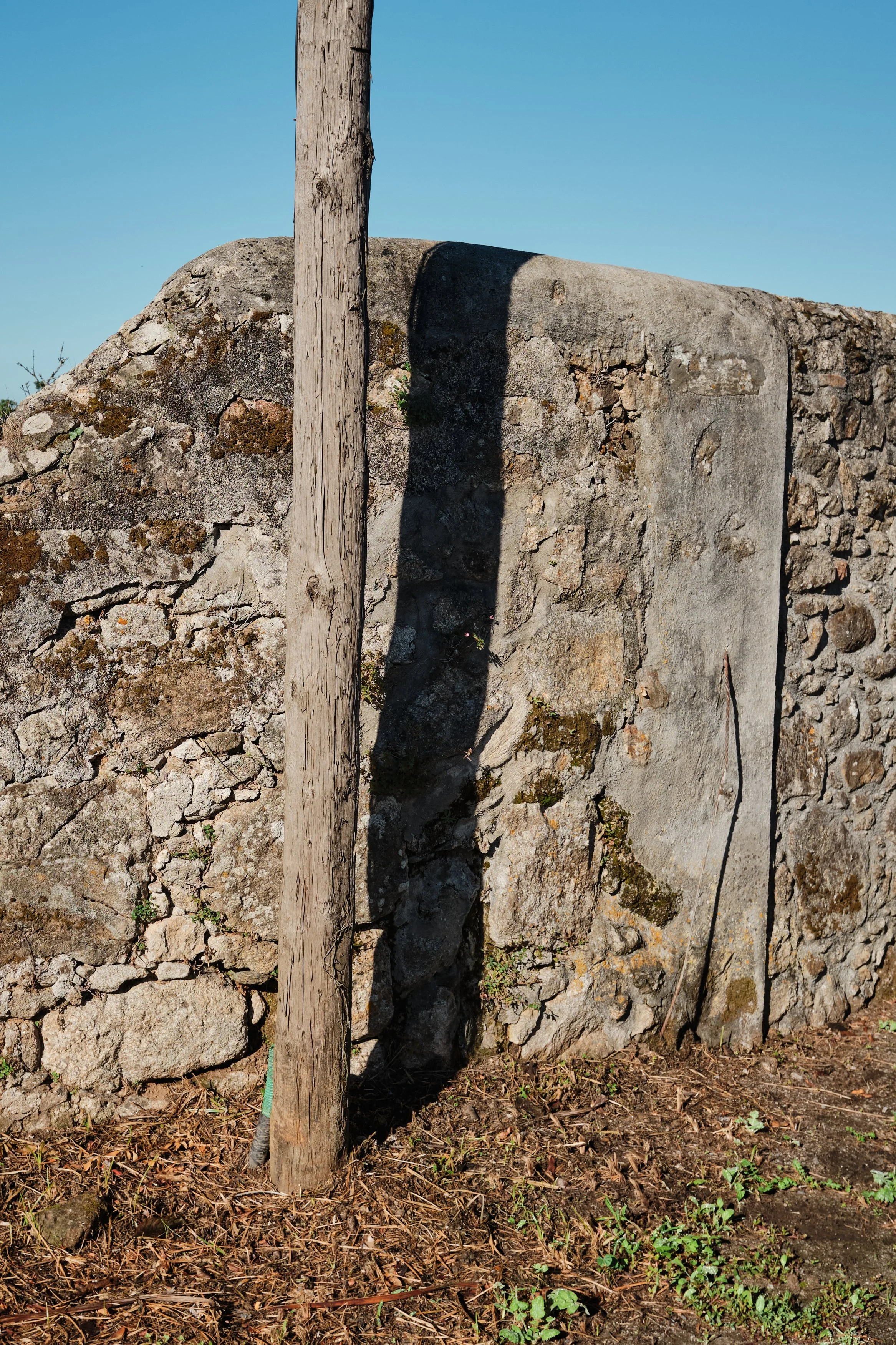 A wooden post standing next to a stone wall with a shadow cast on it from the post under a clear blue sky.