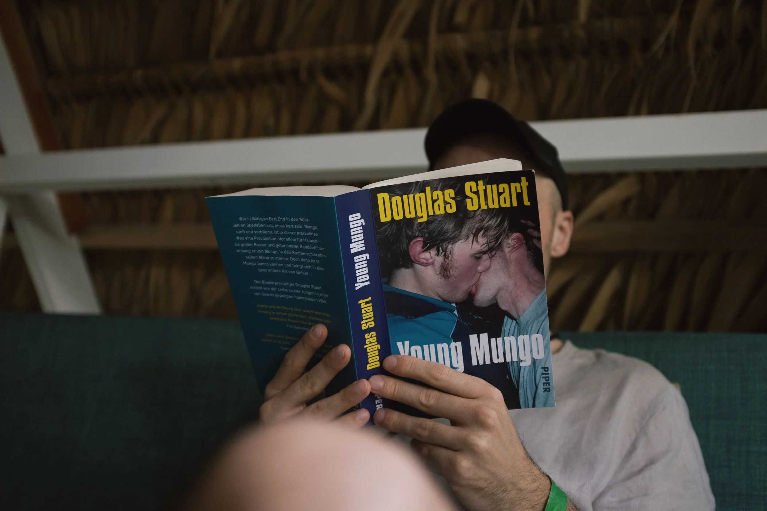 Person sitting on a green couch reading the book 'Young Mungo' by Douglas Stuart. The person is wearing a cap and a light-colored shirt. The background shows a wooden ceiling.