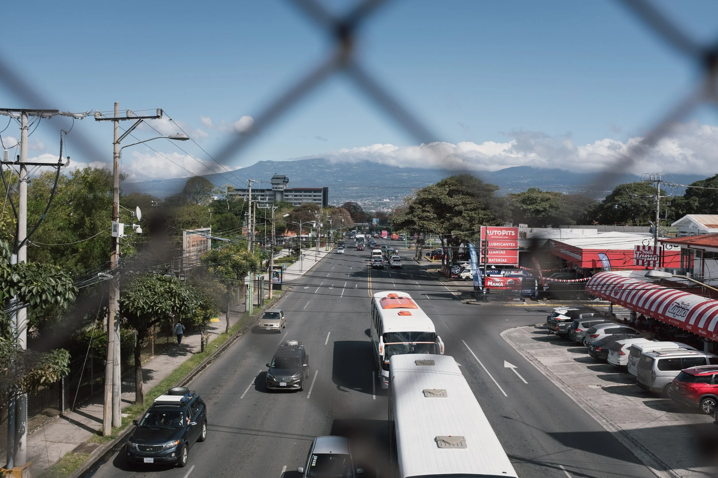 View of a city street seen through a chain-link fence, with cars and buses on the road, shops on the right, and mountains in the background, under a partly cloudy sky.
