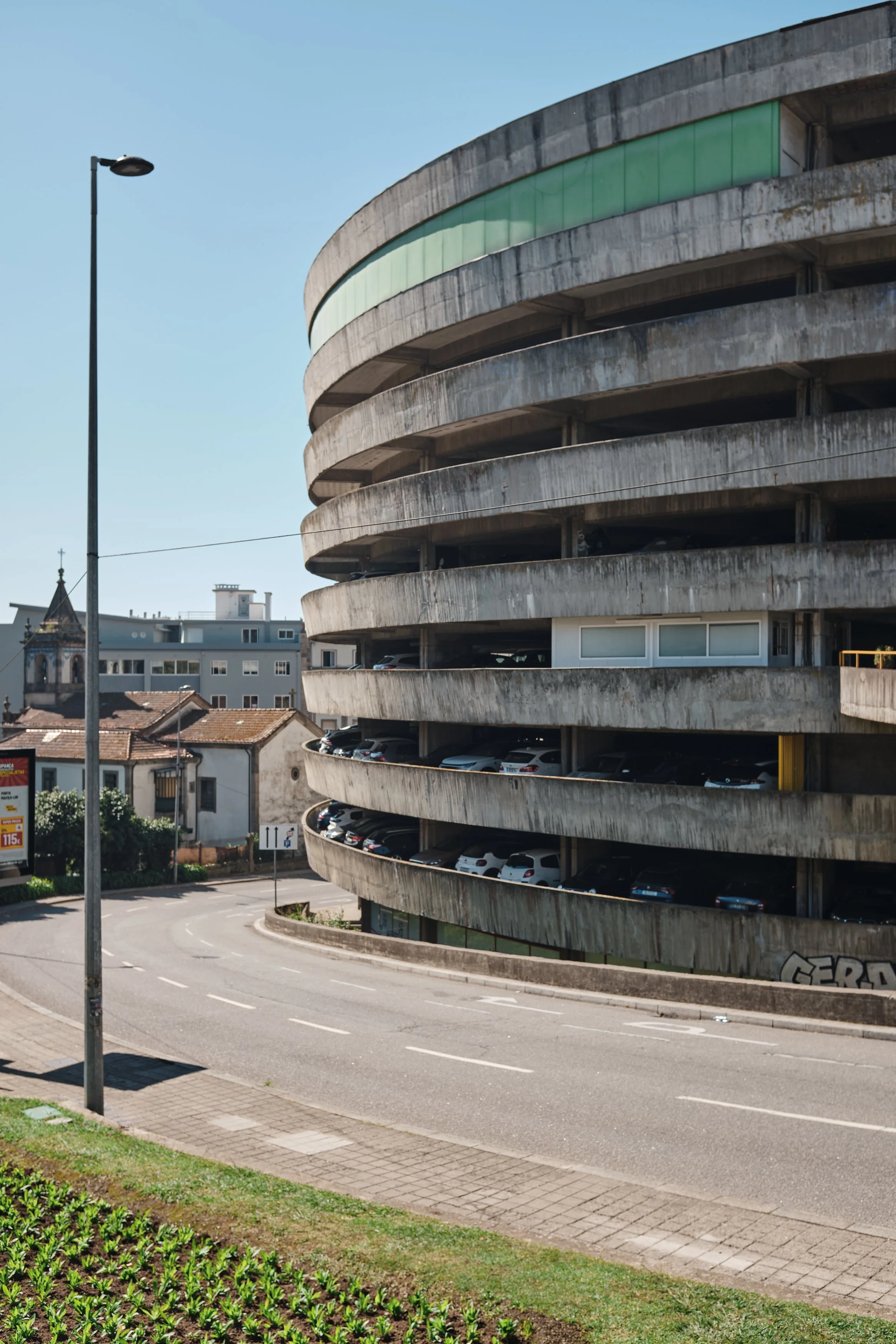 An outdoor parking garage with multiple levels made of concrete, with cars parked inside. Surrounding buildings and a street curve with a sidewalk and a lamppost are also visible under a clear, blue sky.